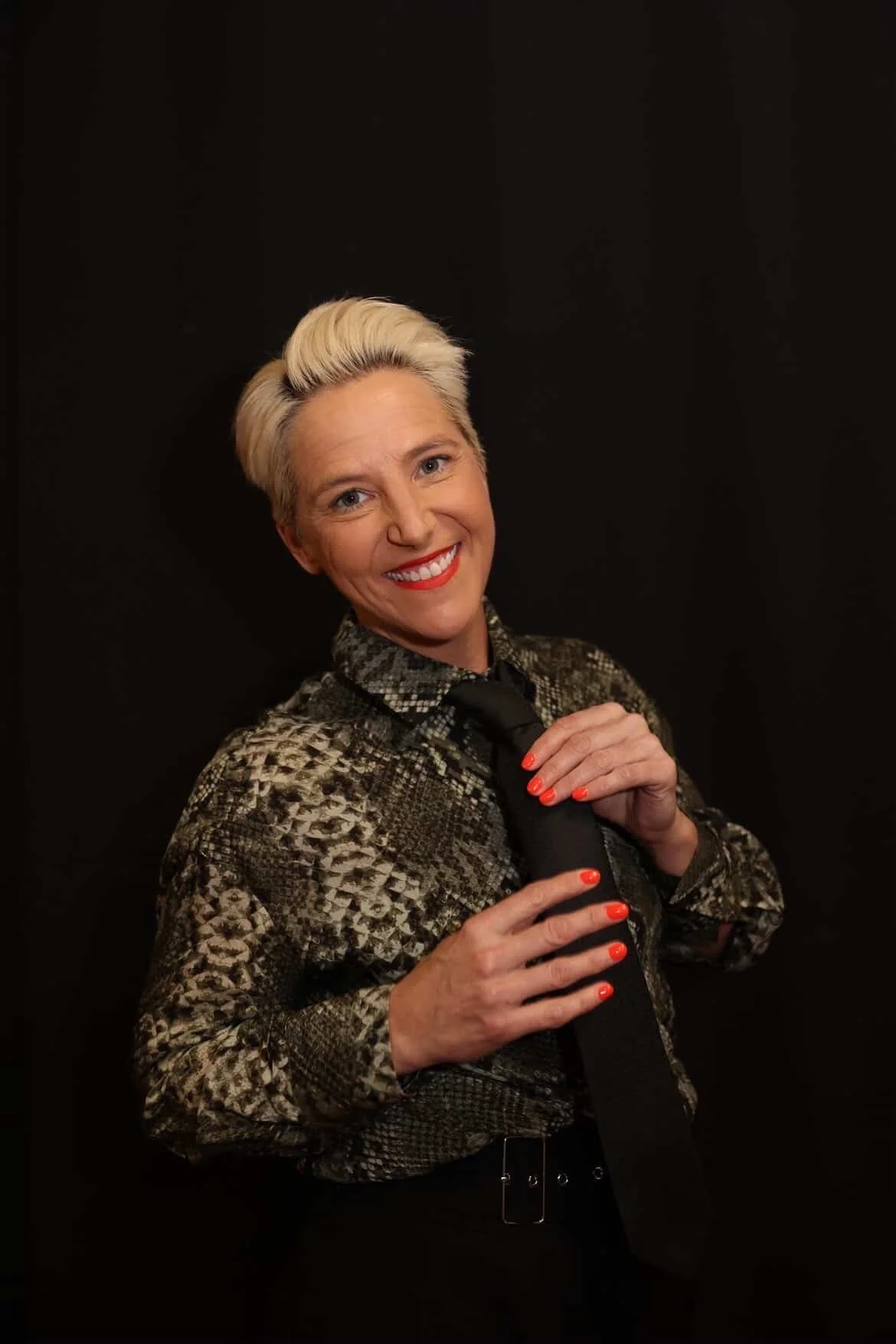 Brooke Weisbord with short, platinum blonde hair and red lipstick, smiling and holding a black tie against a plain black background. She is wearing a patterned blouse and bright orange nail polish.