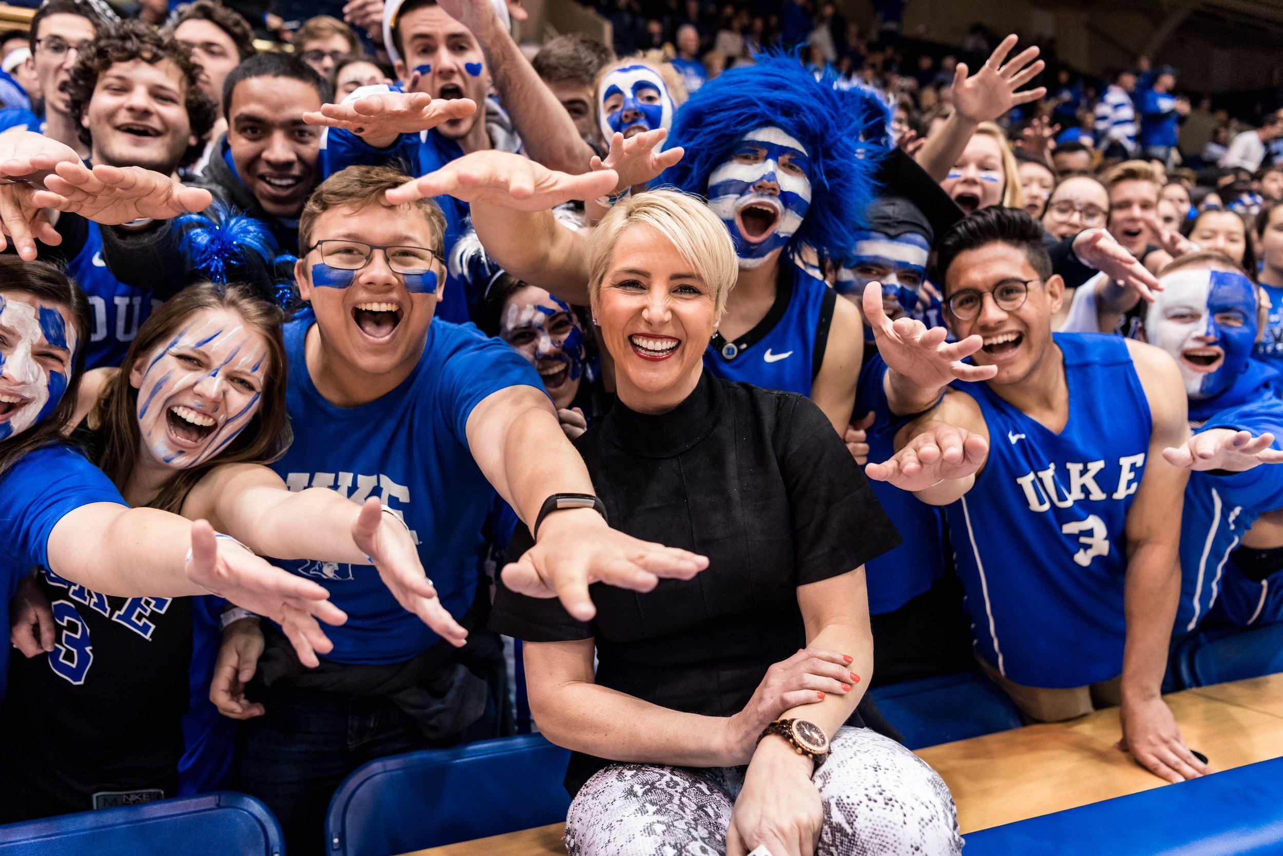 Group of excited sports fans and Brooke Weisbrod in black with short blonde hair at a sports event, wearing blue and face paint supporting Duke University, cheering enthusiastically with arms outstretched.