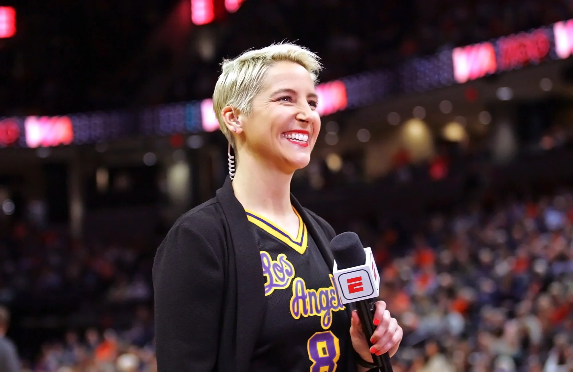 A woman with short blonde hair, wearing a black jersey with yellow and purple details, smiling and holding a microphone during an interview at a sports arena.