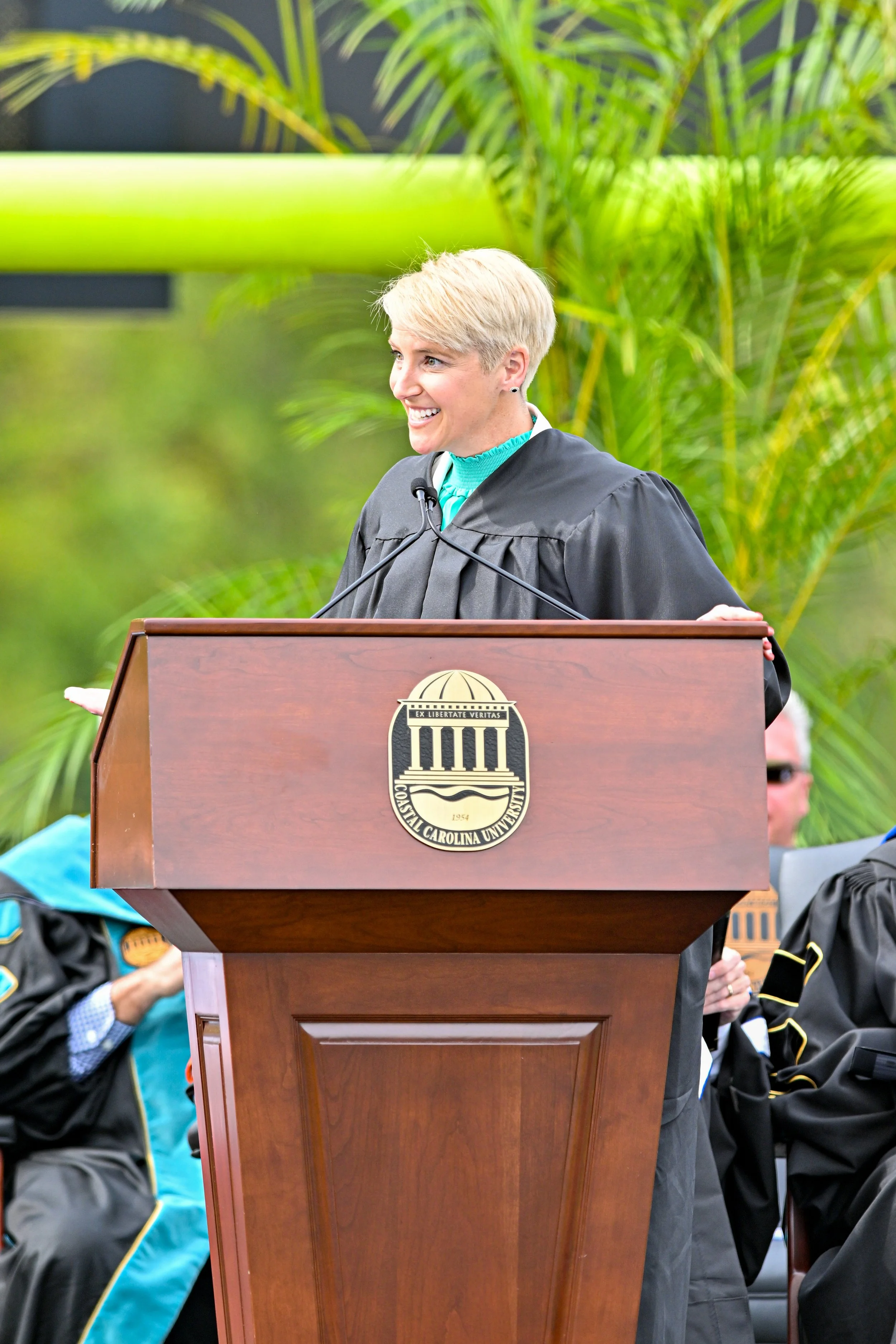 A woman with short blonde hair smiling while speaking at a podium with the Coastal Carolina University logo, with several people seated nearby and green foliage in the background.