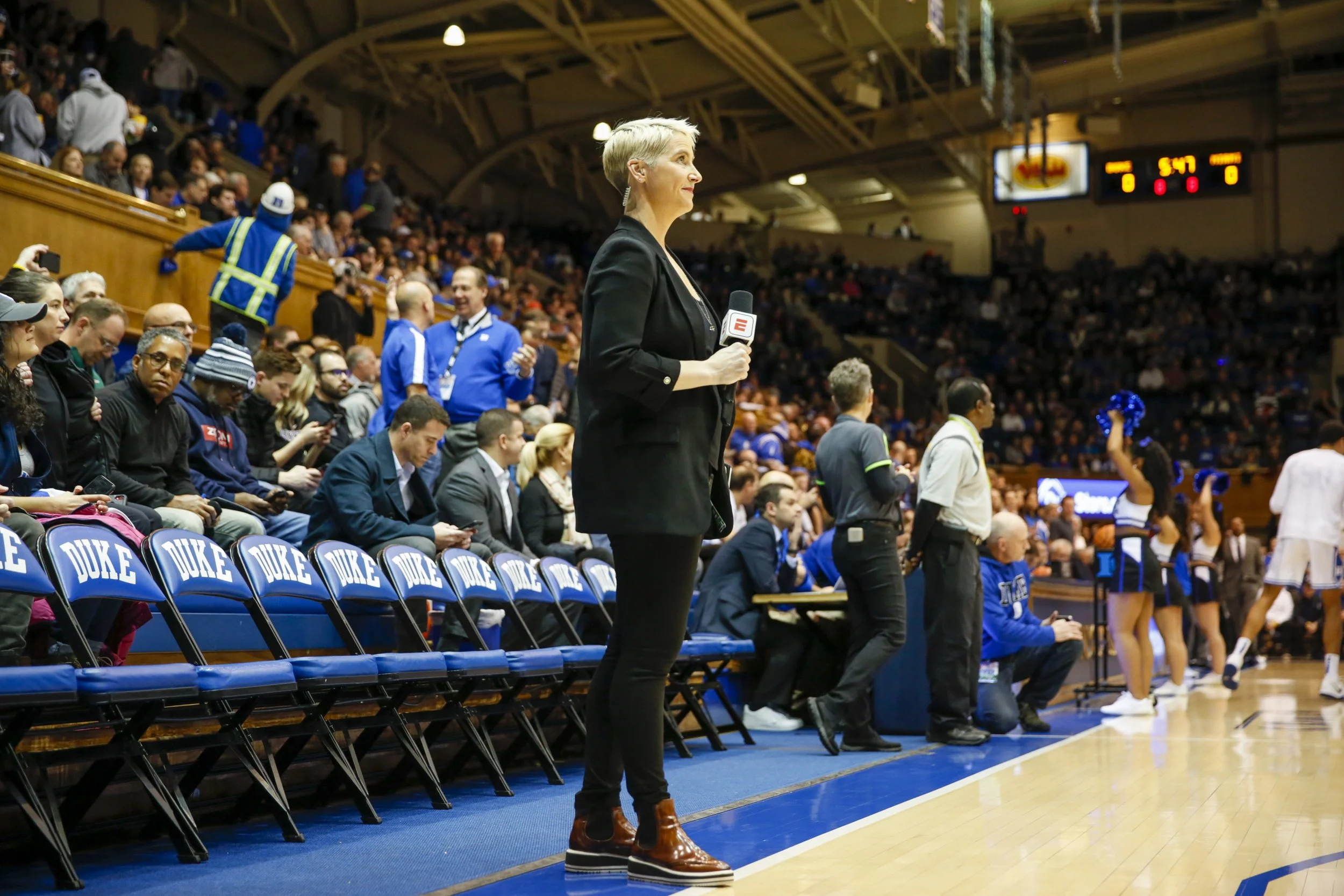 A woman stands courtside at a basketball game, holding a microphone with the ESPN logo. She is dressed in a black blazer, black pants, and brown boots. The stands behind her are filled with spectators, some of whom are using their phones. The row of seats in front of her has blue chairs with