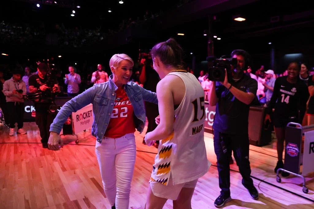 Two women are dancing and smiling on a basketball court, surrounded by a crowd and a cameraman capturing the moment. The woman on the left has short blonde hair, wears a denim jacket and a red basketball jersey with the number 32. The woman on the right has her hair tied back, wears a white basketball jersey with the number 15, and beige shorts.