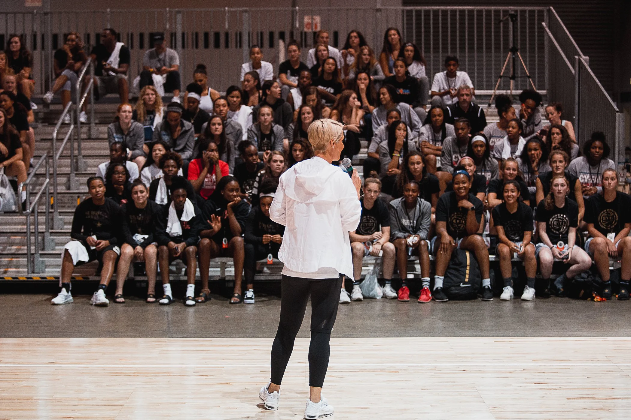 A person with short blond hair, wearing a white shirt and black leggings, holding a microphone, standing in front of an audience of mostly young women seated on bleachers in an indoor gymnasium.