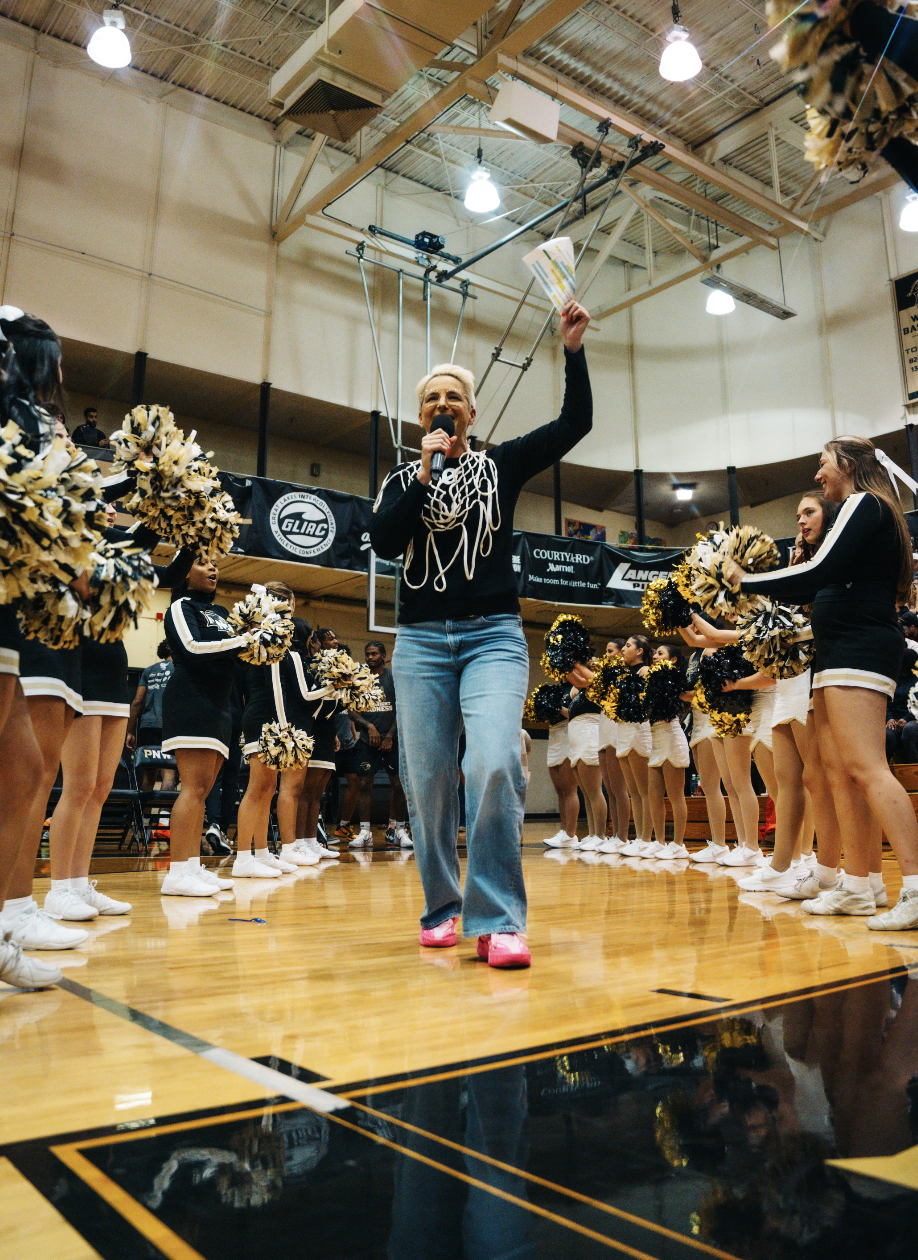 A woman with short blonde hair holding a microphone and raising her hand holding papers, leading a cheer squad in a basketball gymnasium during a cheerleading event. The cheerleaders are in black and white uniforms with yellow and black pom-poms, standing in formation on the gym floor.