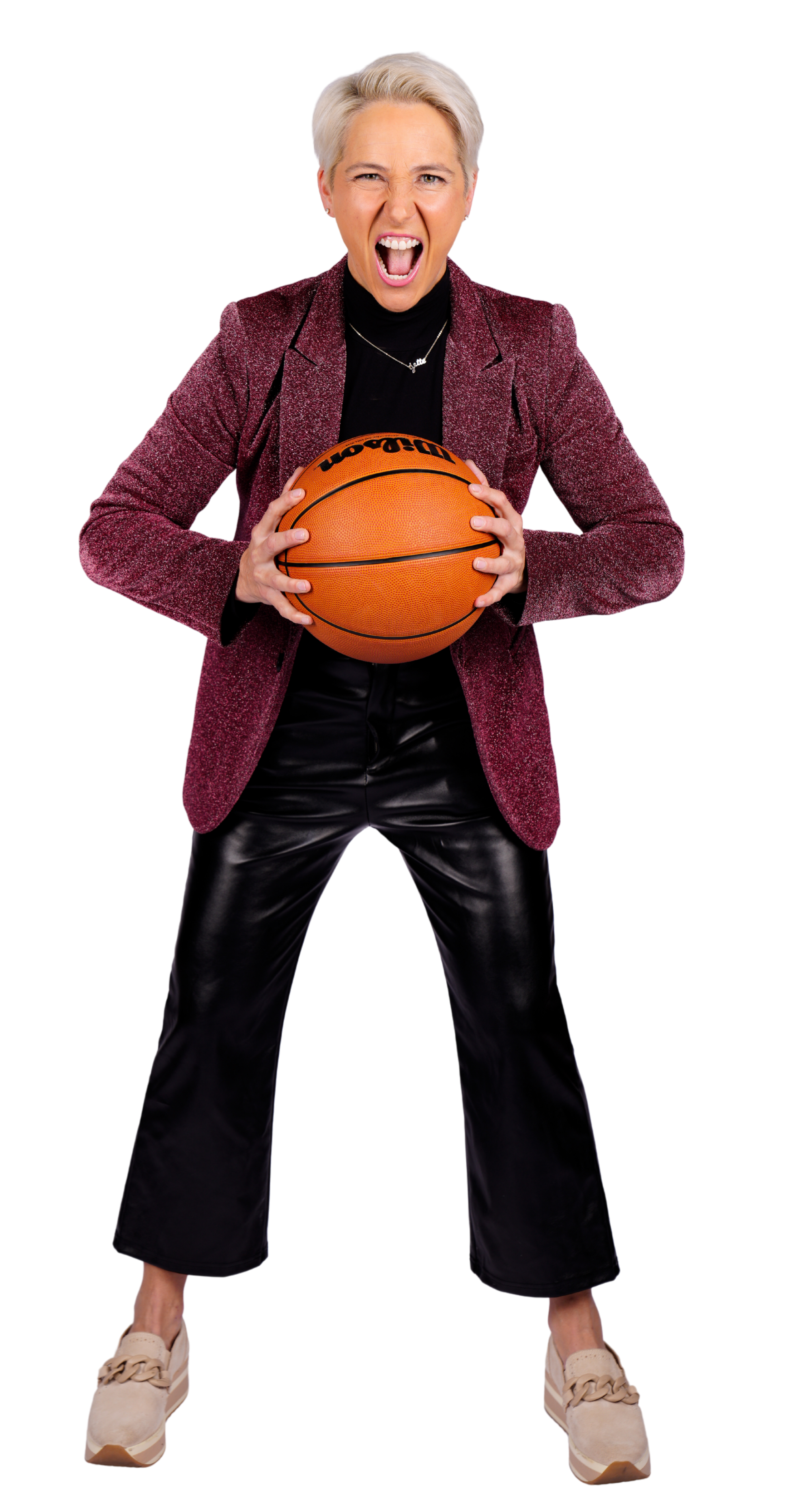 A woman with short gray hair wearing black leather pants, a burgundy blazer, and beige sneakers, holding a basketball and shouting.