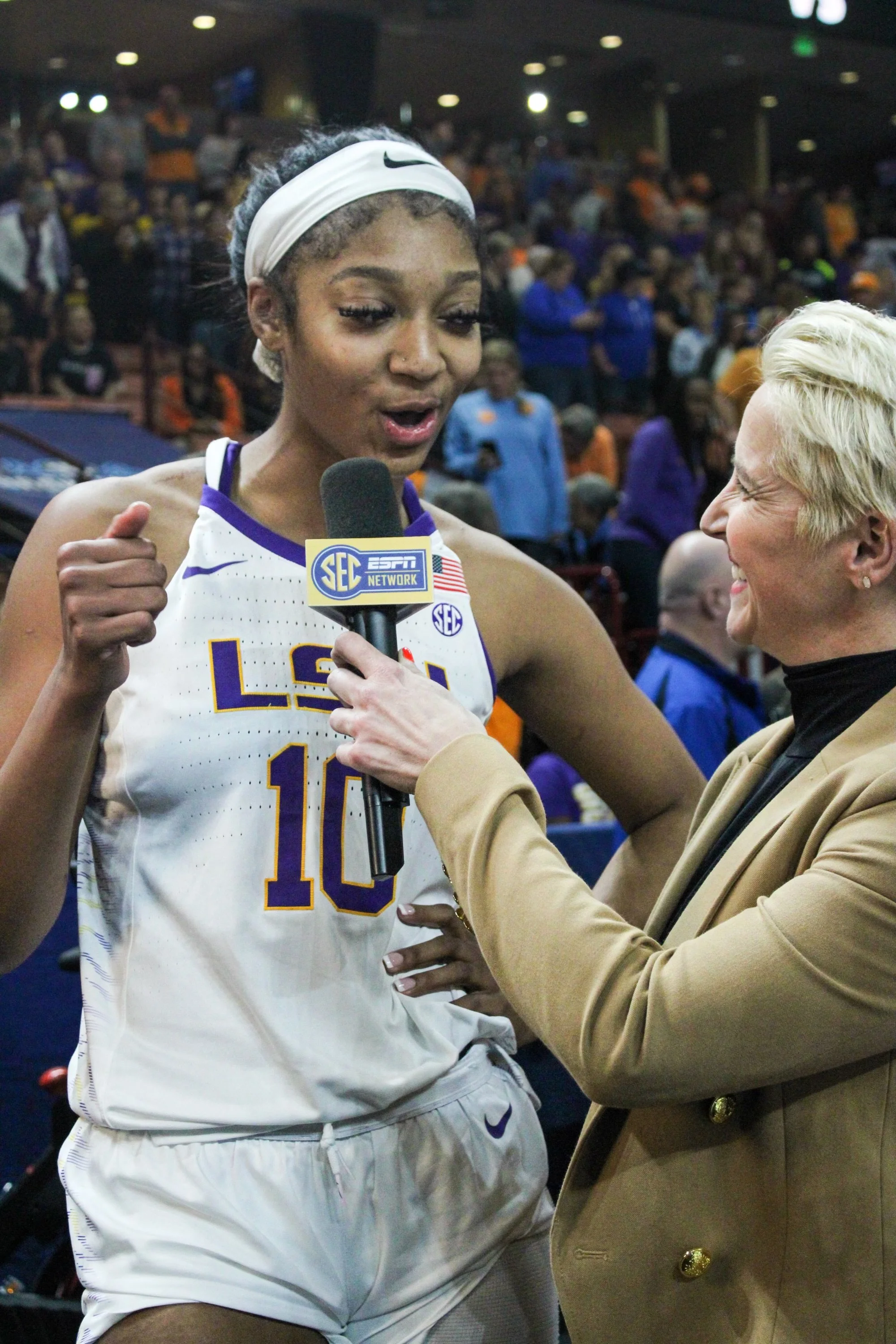 A female athlete being interviewed after a basketball game, wearing a white LSU jersey with the number 10, holding a microphone with the SEC ESPN Network logo, standing in a crowded sports arena.
