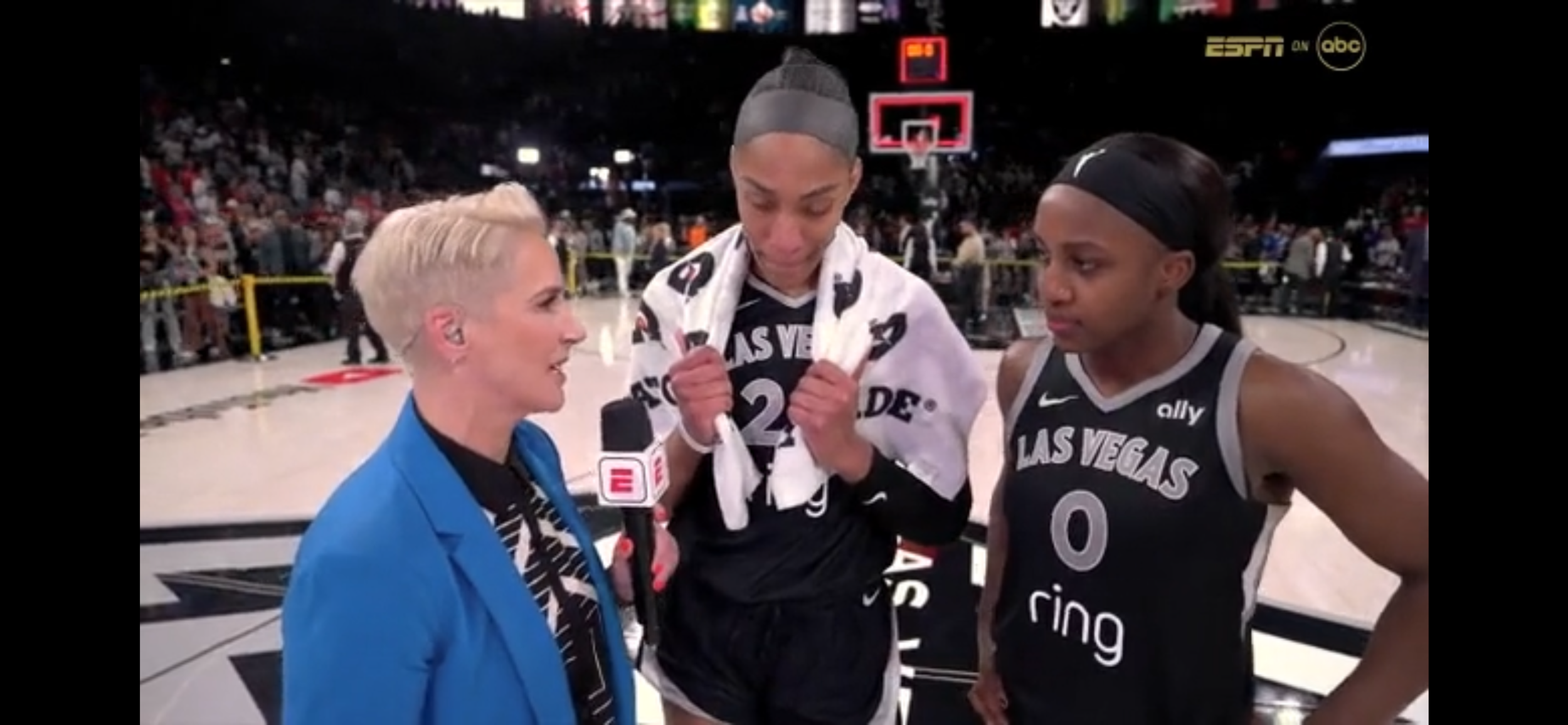 Three women basketball players wearing black jerseys with 'Las Vegas' on them are interviewed by a woman in a blue jacket after a game. The woman in blue is speaking into an ESPN microphone. The players are holding their towels and are standing on the basketball court in a stadium filled with crowd.
