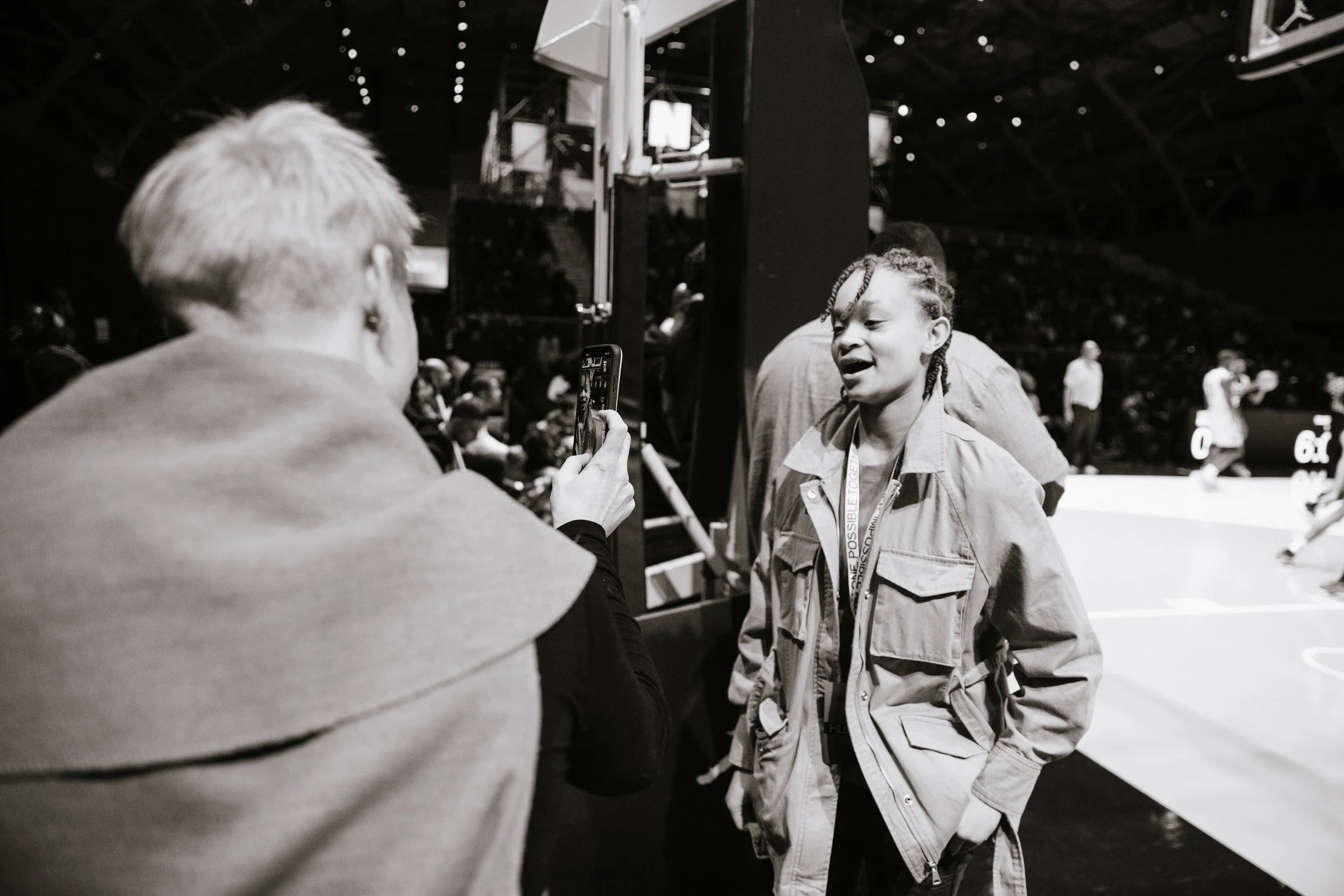 A young person with braided hair and a jacket talking to an older man with short hair, who is taking a photo of them with a smartphone at an indoor sports event.