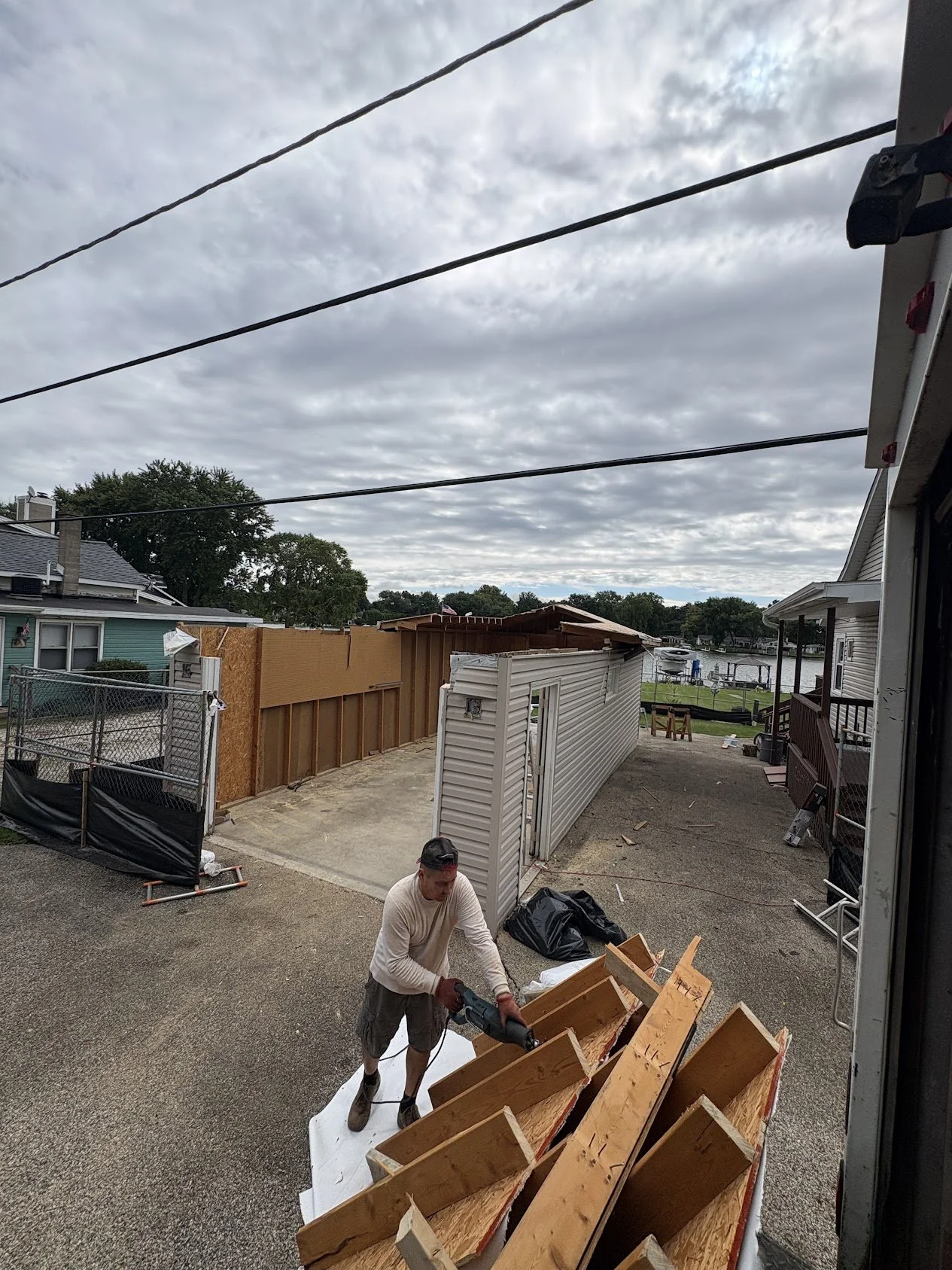 Man working outdoors on construction or renovation project, handling wooden furniture or cabinets, with a house, a partially built shed, and a waterway in the background.