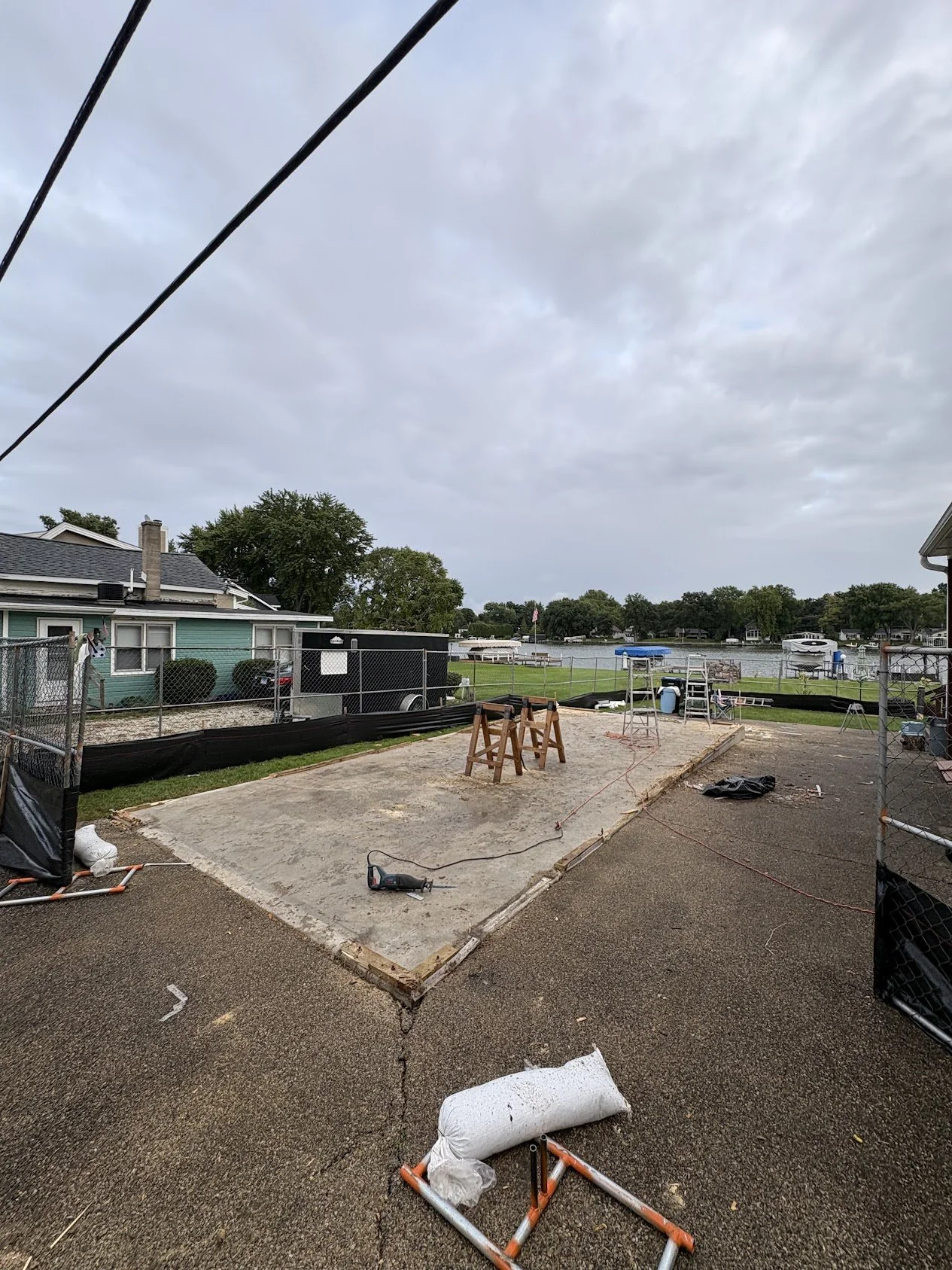 Backyard construction site with a concrete slab, wooden frames, and tools, overlooking a lake with boats, trees, and houses under an overcast sky.