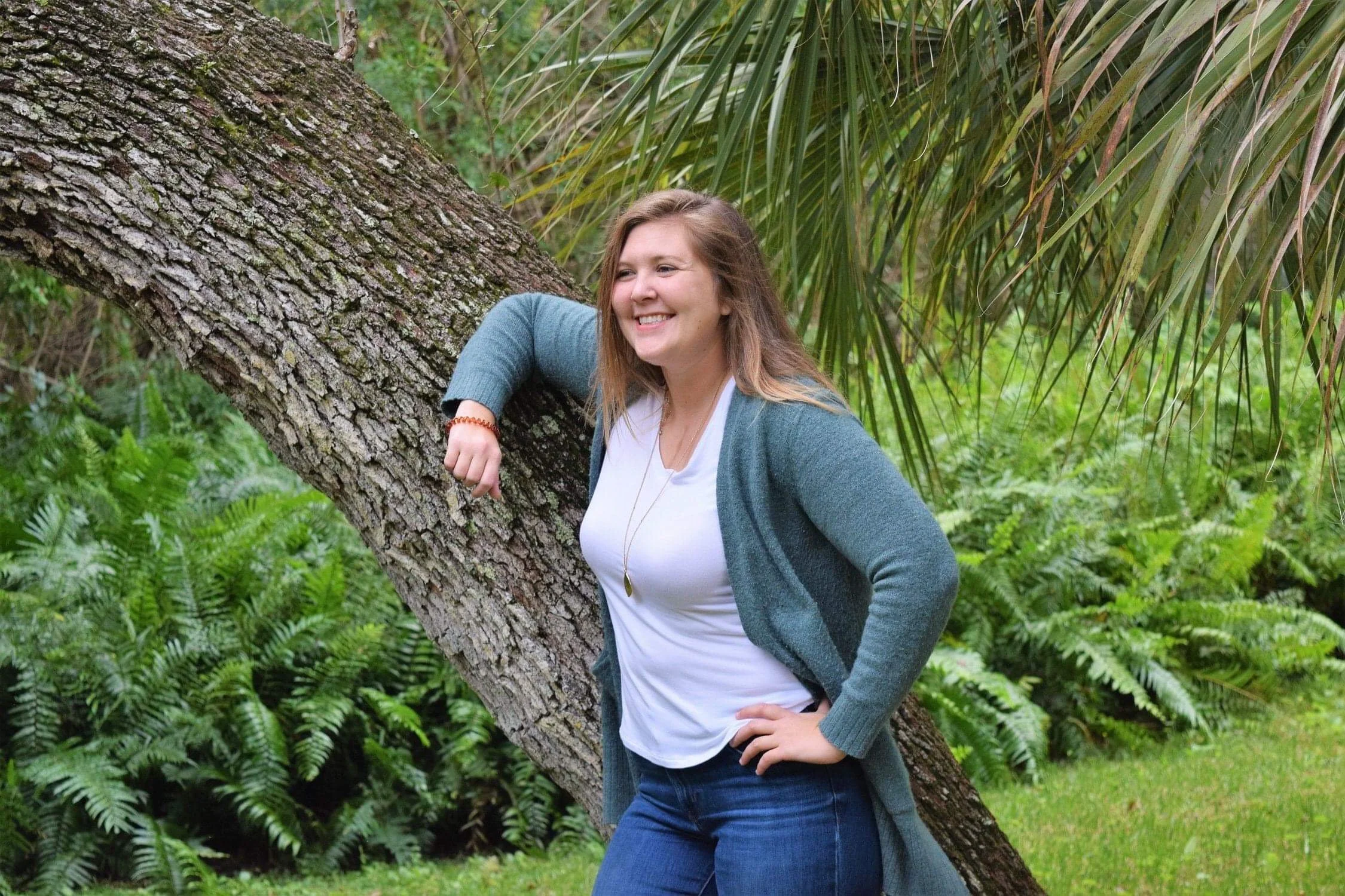 Teen girl smiling and leaning against a tilted tree trunk in a lush green forest.