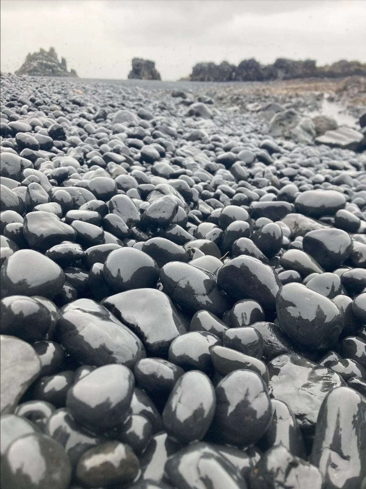Close-up of wet, black, and gray pebbles on a beach with rocks and cliffs in the background under an overcast sky.