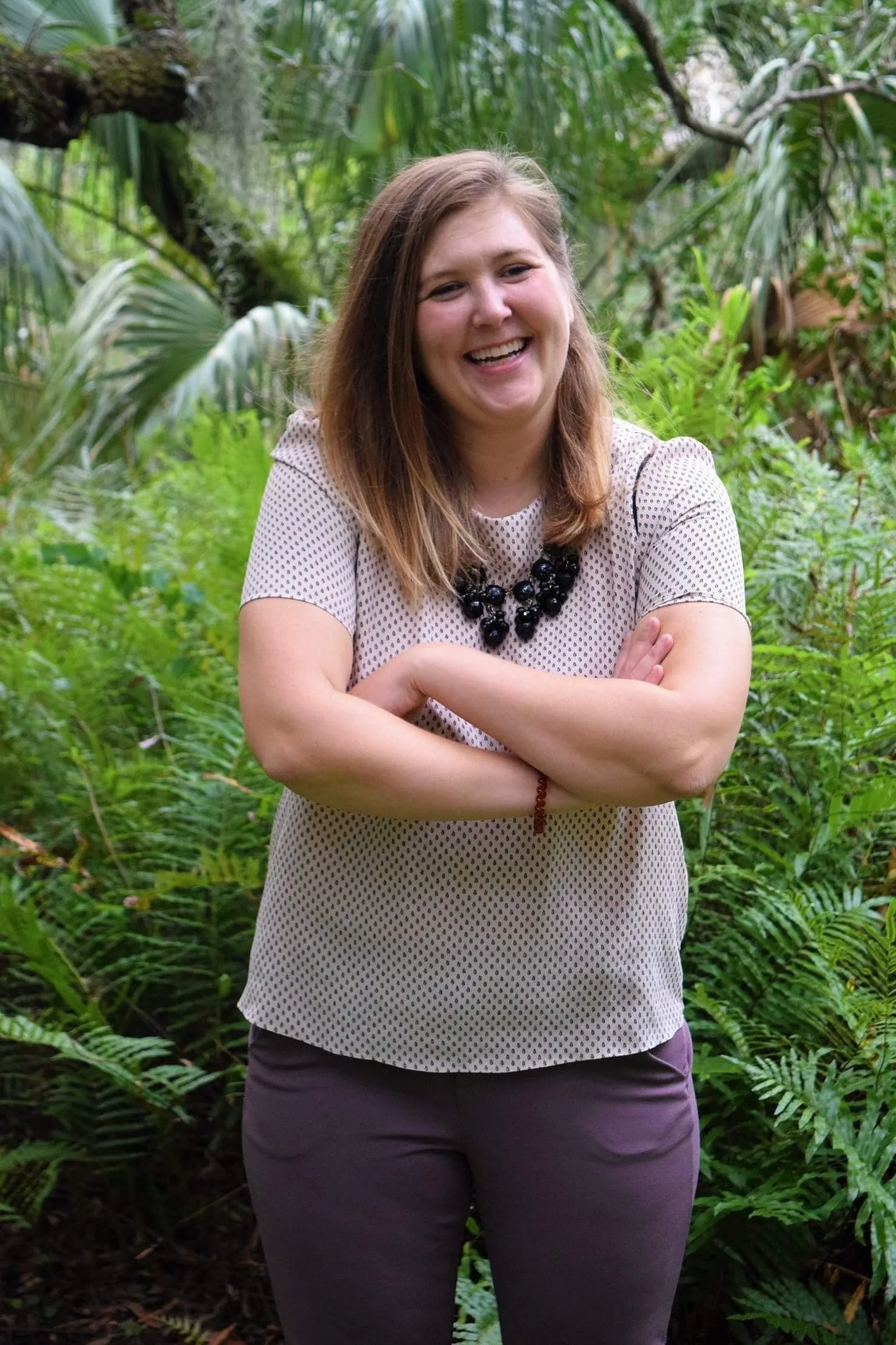 A woman with light brown hair wearing a white dotted blouse and dark pants, smiling and crossing her arms in front of lush green tropical plants.