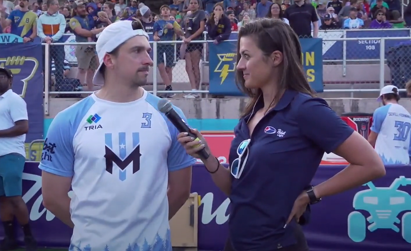 A woman with a microphone interviewing a man in a sports stadium with spectators in the background. The man is wearing a white and light blue sports jersey with the number 3, and the woman is wearing a dark blue shirt with sunglasses hanging from her collar.