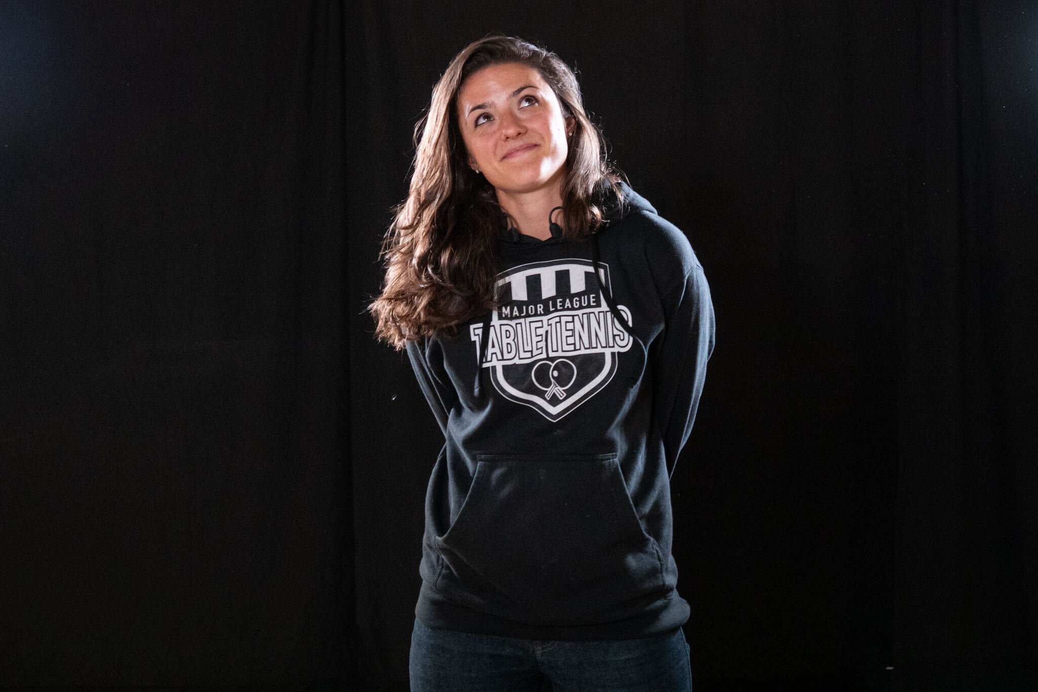 A young woman with long brown hair wearing a black hoodie with a ping-pong logo, standing against a black background, looking up and smiling.