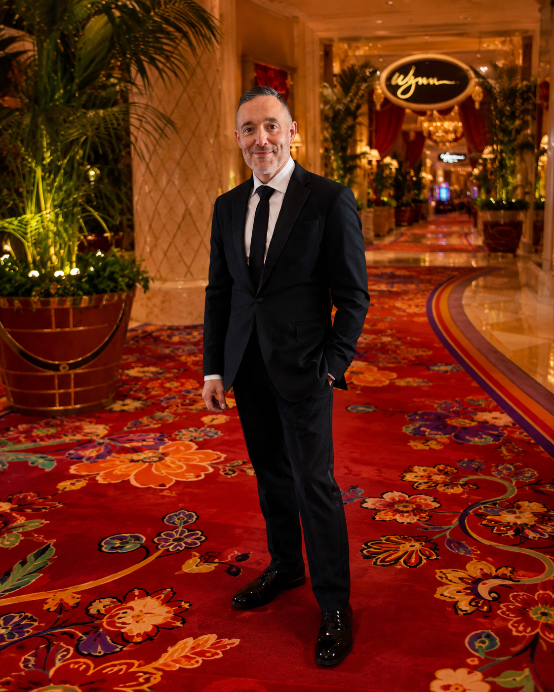 A man in a black suit and tie standing in The Wynn with red floral carpet, large potted plants, and warm lighting.