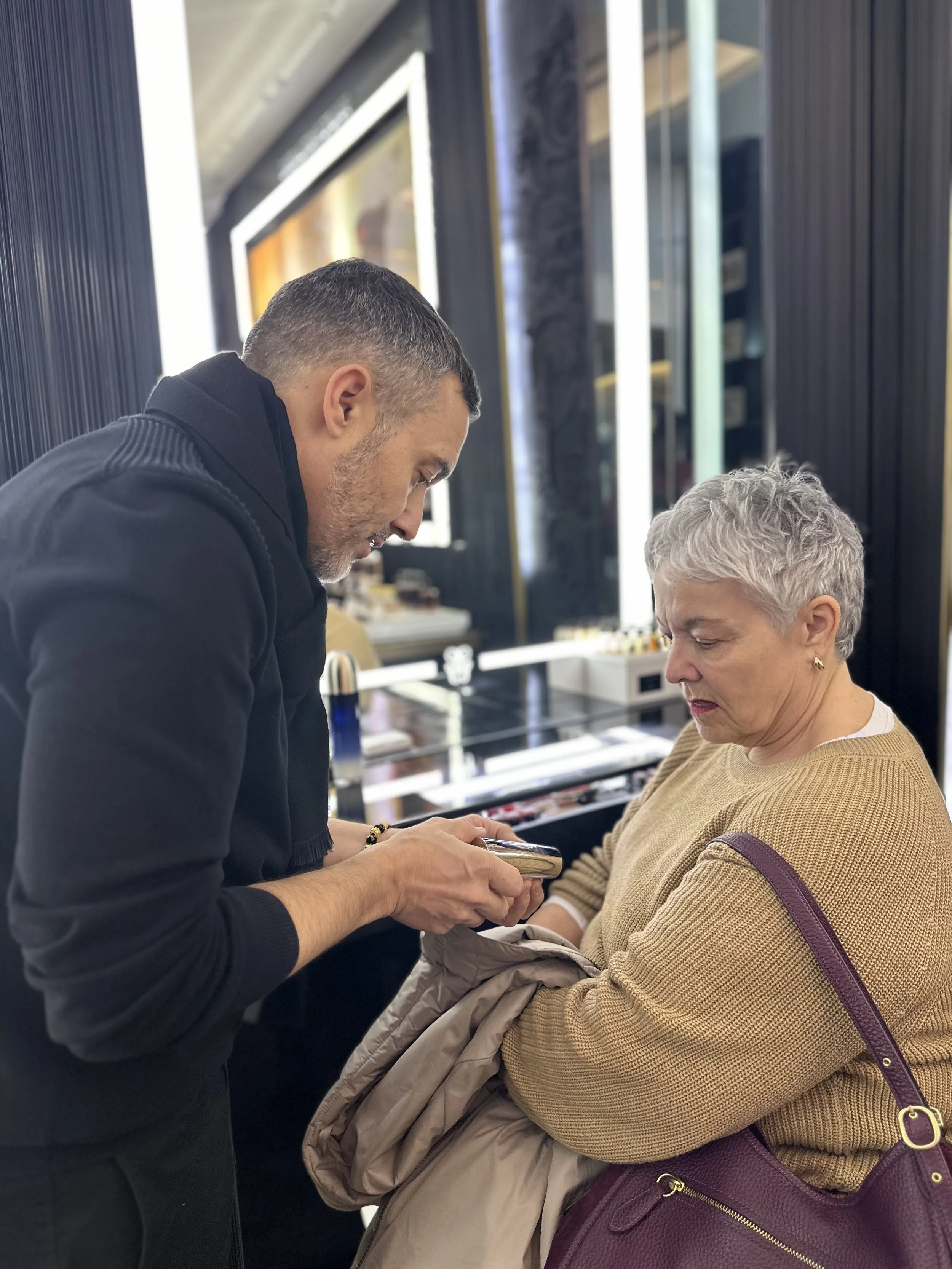 A man with short dark hair and a beard showing a woman with short light gray hair sharing a bottle of serum, inside a boutique with dark wood and black walls.