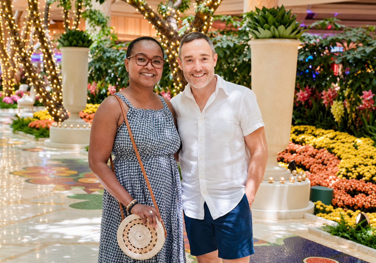 A smiling couple standing indoors among bright, colorful floral arrangements and decorative lights, with large potted plants and patterned floor.