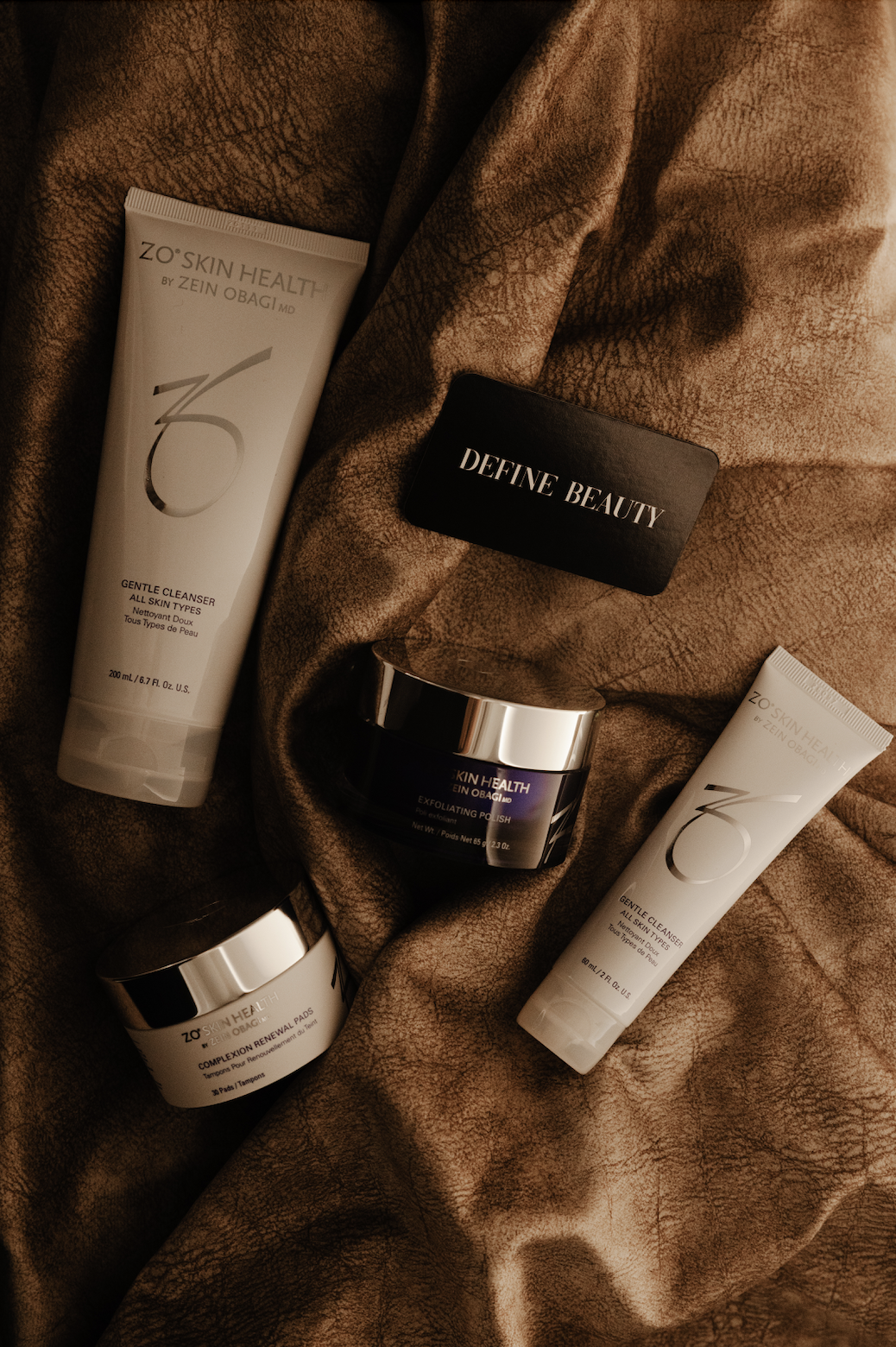 Skincare products including two tubes of ZO skin health gentle cleanser, two jars of ZO skin health exfoliating polish and renewal pads, and a black box labeled 'Define Beauty' arranged on a textured brown fabric background.