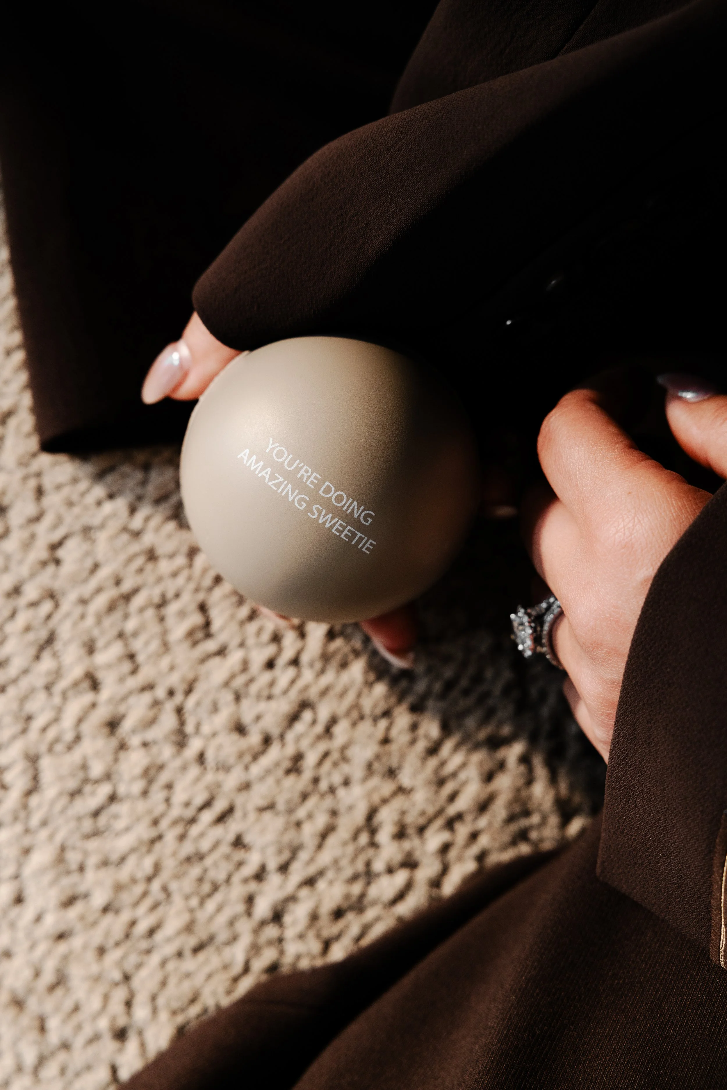 Close-up of a person's hand holding a spherical object with the text 'You're doing amazing sweetie' printed on it, on a brown fabric background.