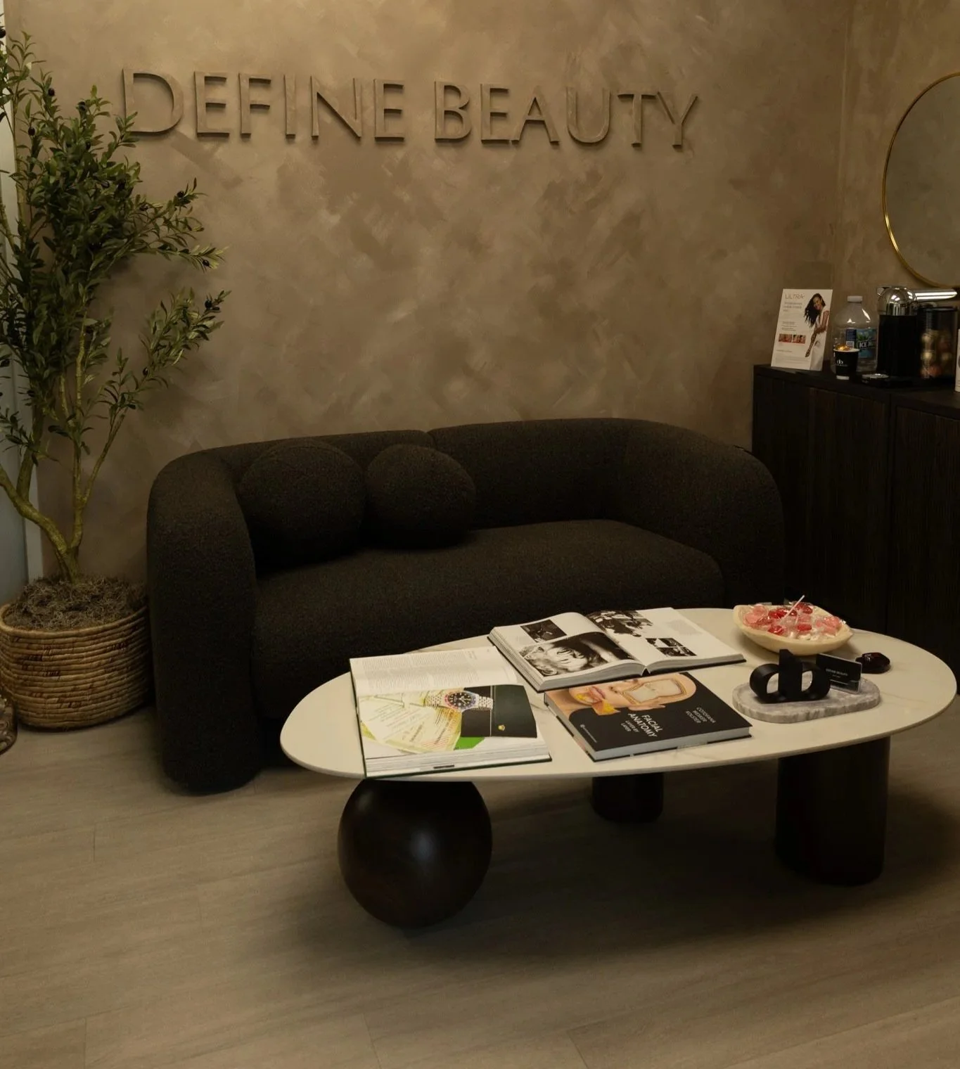 An interior scene of a waiting or lounge area with a black sofa, a white coffee table with magazines and decor, a potted plant on the left, and a wall with the words 'DEFINE BEAUTY' displayed. On the right, a black cabinet holds a water bottle, a coffee machine, and a framed picture, with a small round mirror above.