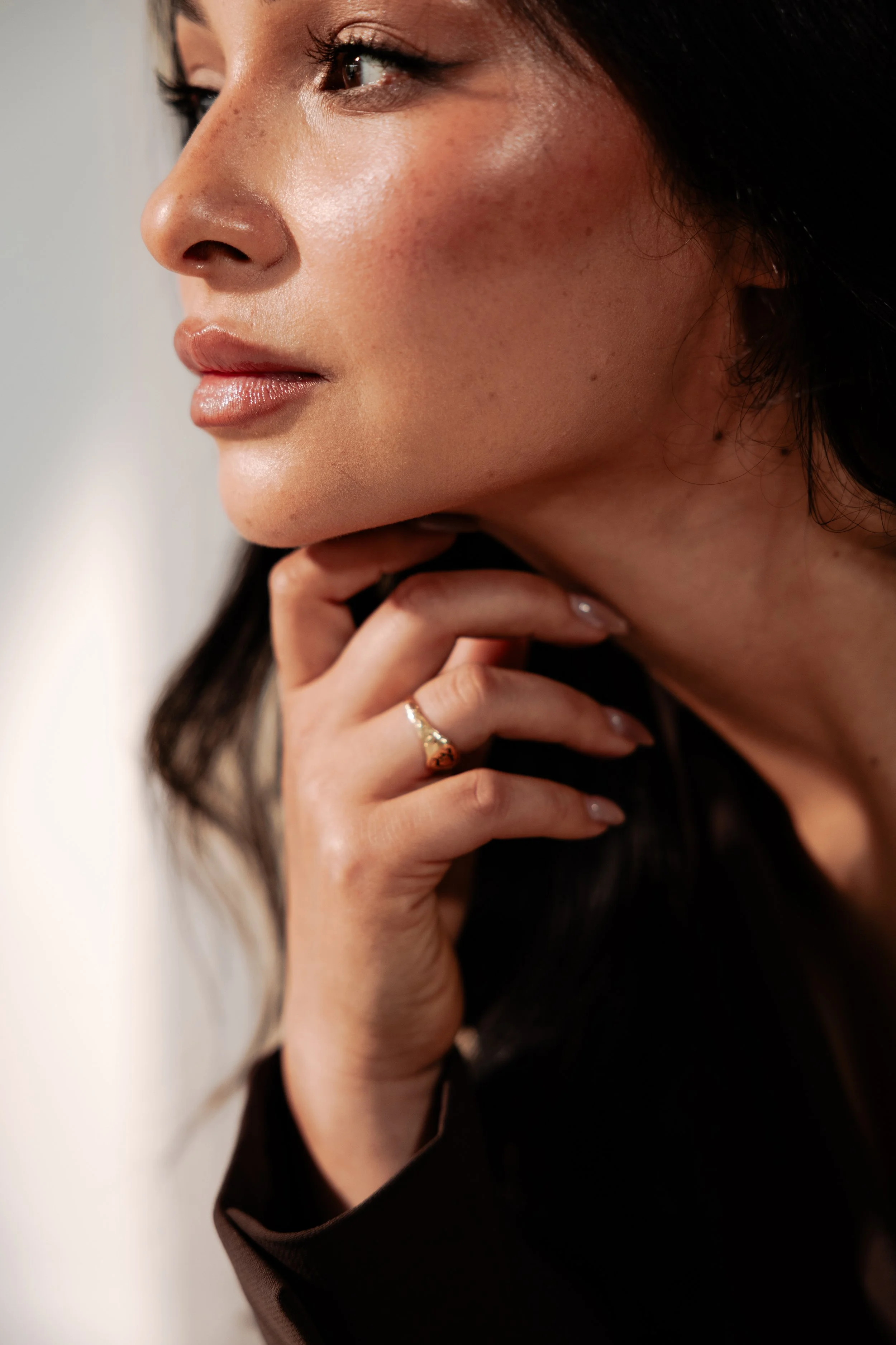 Close-up of a woman with long dark hair, looking to the side, with her hand gently resting near her chin. She has smooth skin, subtle makeup, and wears a ring on her finger.