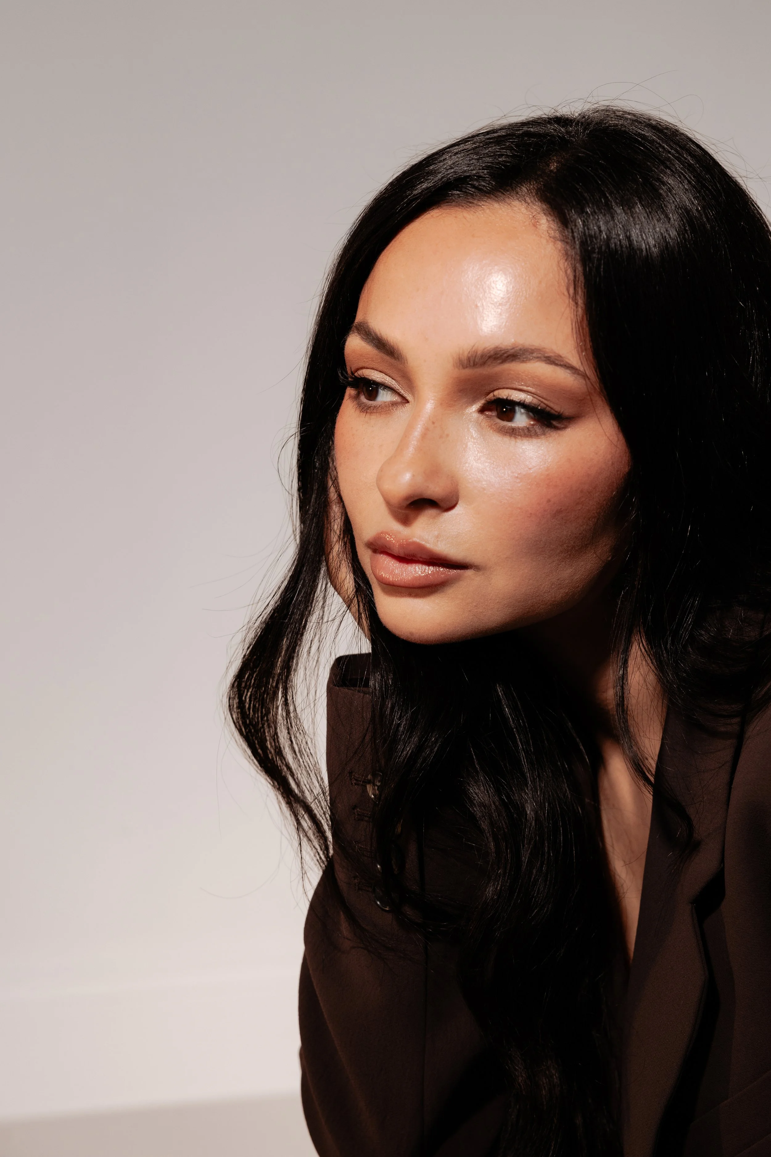 Close-up of a woman with long black hair, wearing makeup, and a dark brown blazer, against a plain light background.