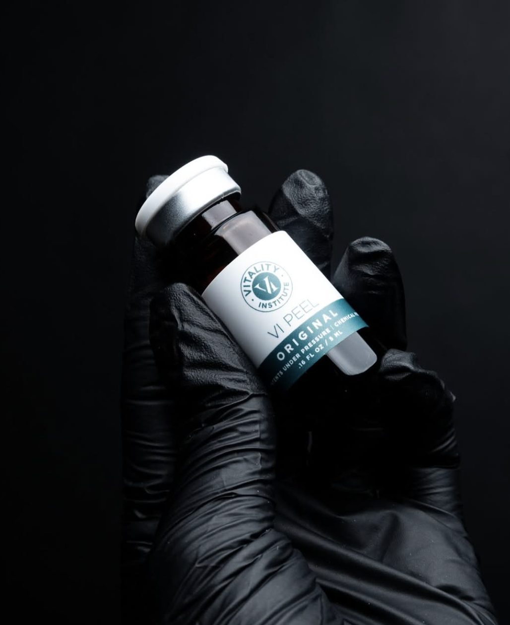 A hand wearing a black glove holding a small brown glass bottle labeled 'Vitality Institute V Peel Original' against a black background.