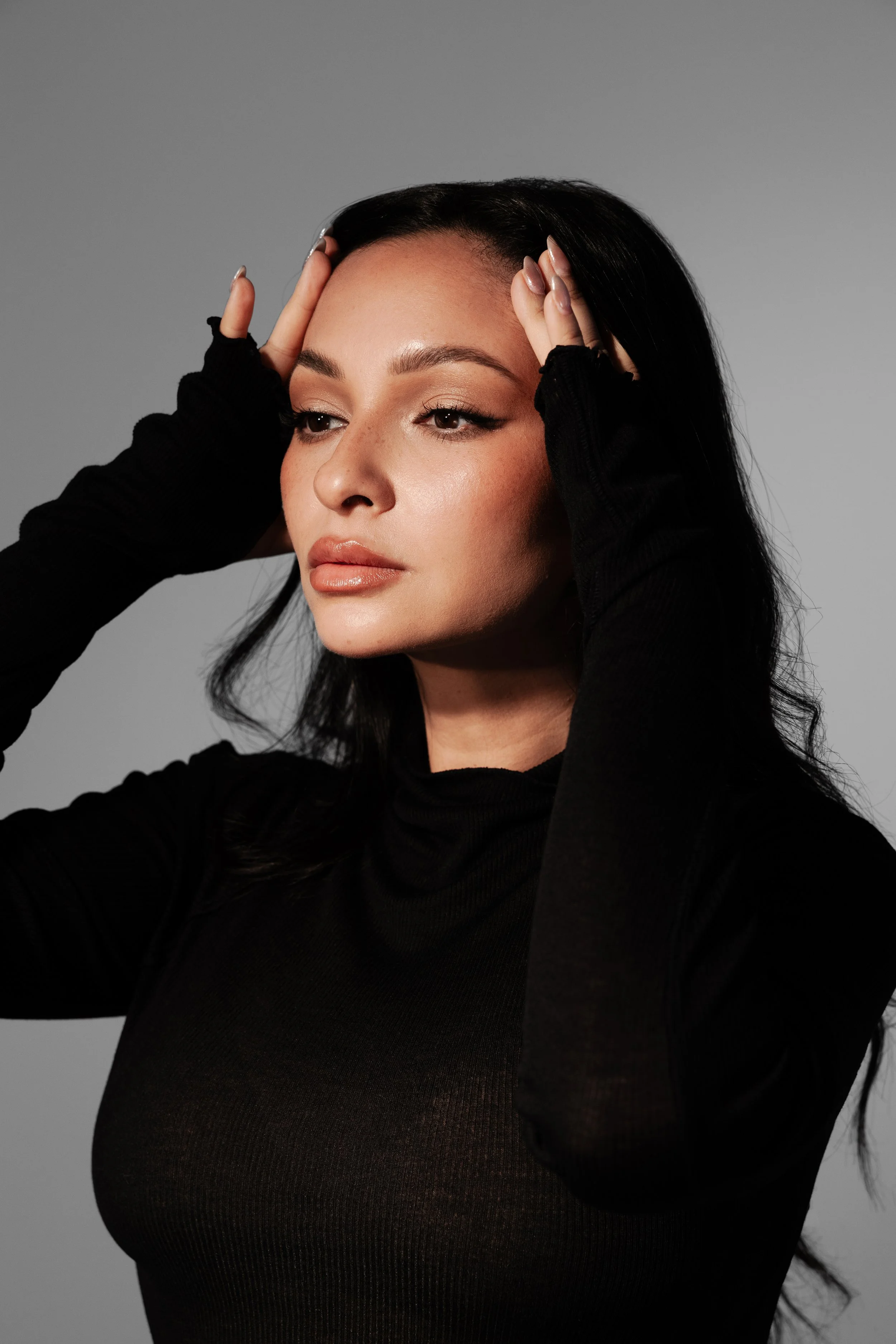 Close-up of a woman with black hair, wearing a black long-sleeve shirt, touching her forehead with both hands, against a gray background.