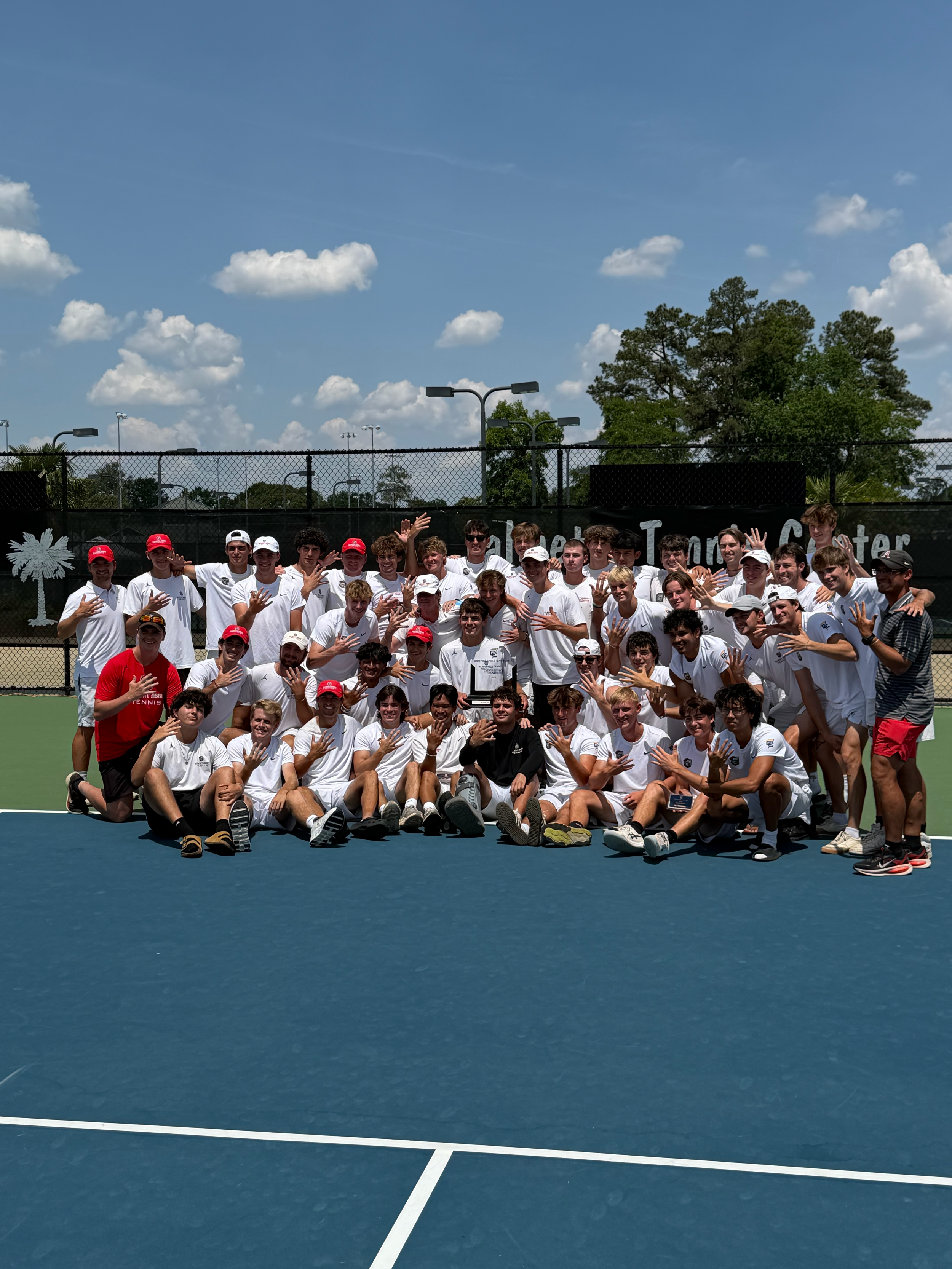Group of young tennis players and coaches in white shirts, some wearing caps, posing for a photo on an outdoor tennis court with blue surface, fence, and cloudy sky in the background.