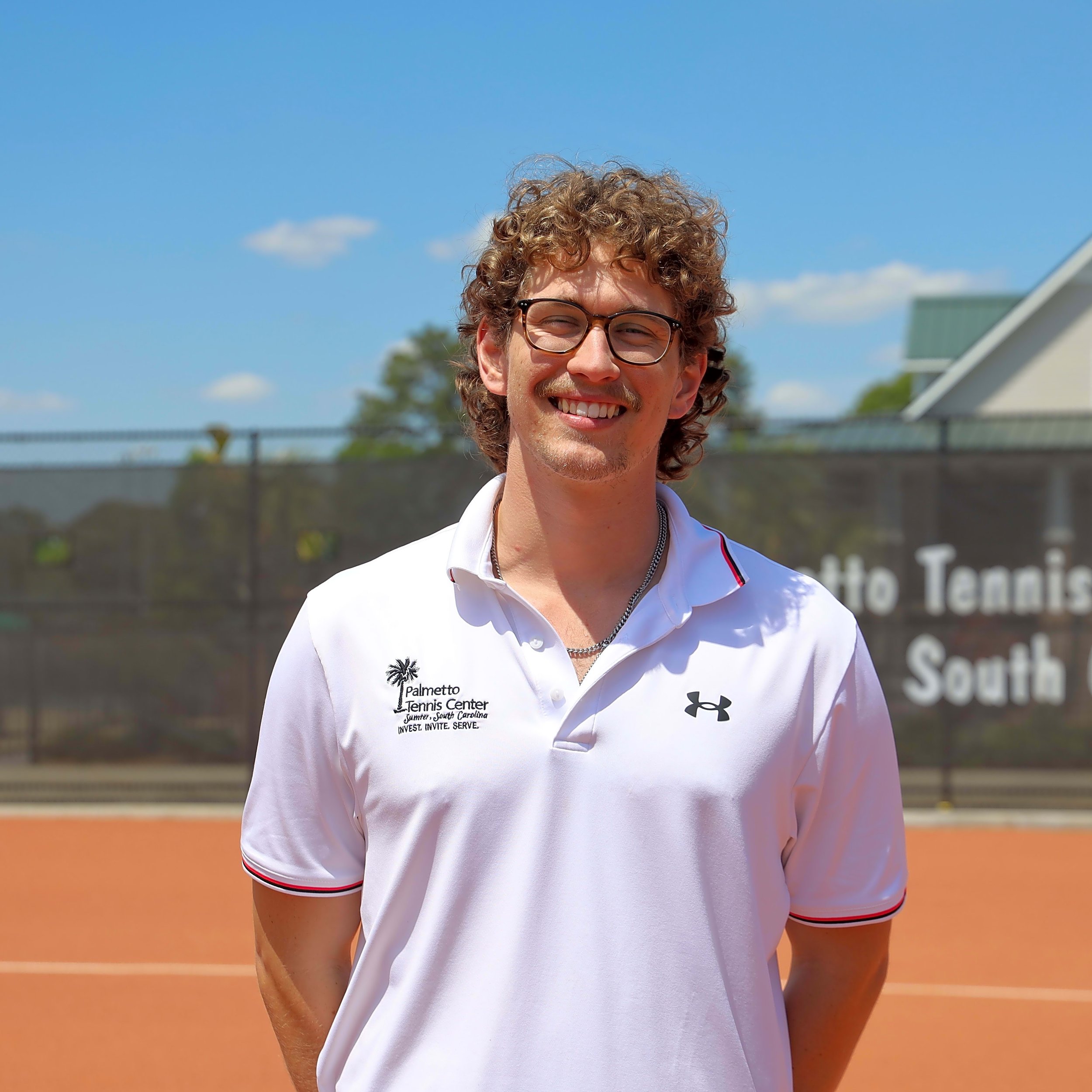 Close-up of a young man with curly hair, glasses, and a big smile, standing outdoors at a tennis court during daytime, wearing a white volleyball shirt with the logo of Palmetto Tennis Center.
