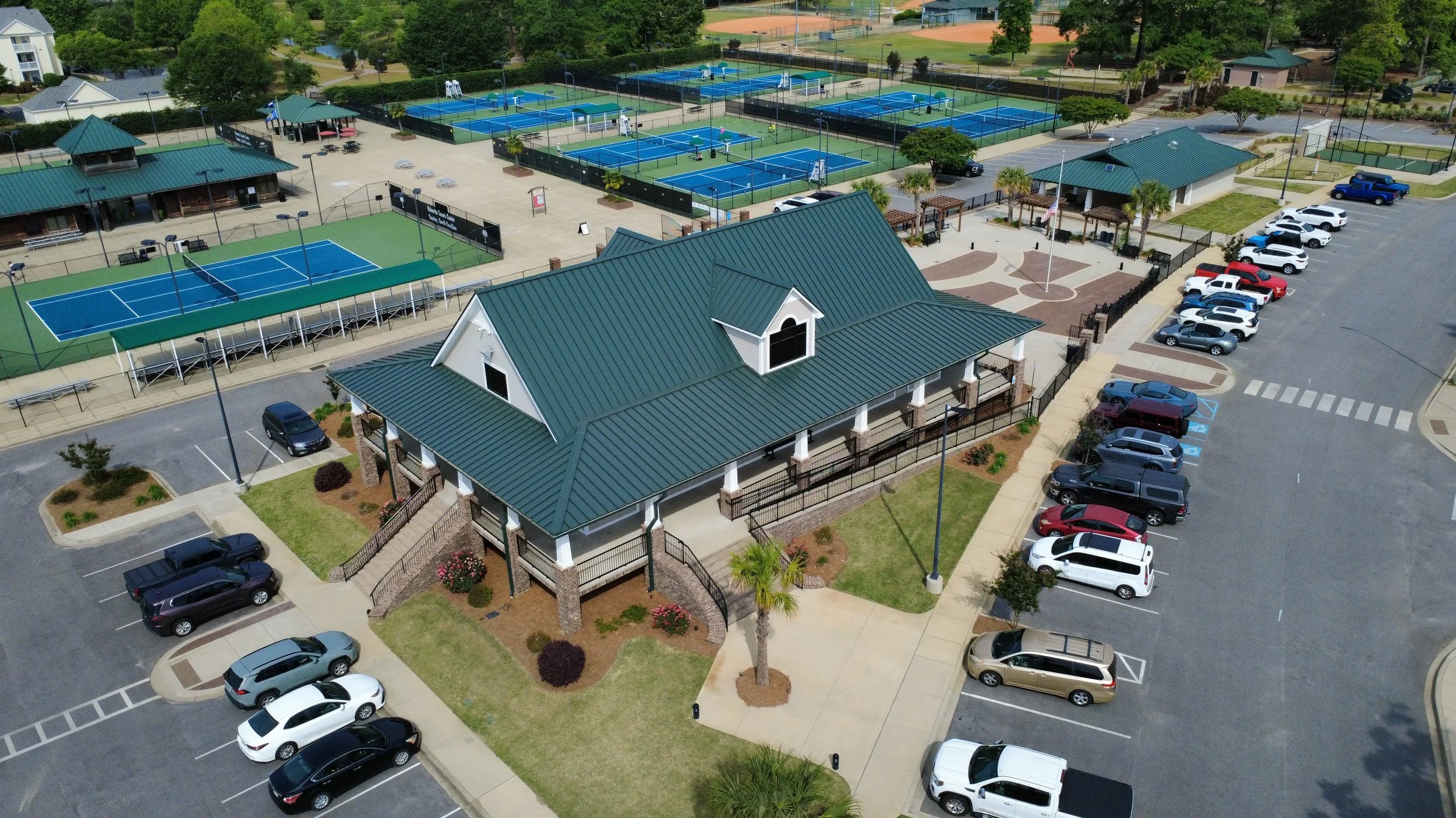 Aerial view of a sports facility with multiple tennis courts, a small building with a green roof, parking lots filled with cars, and some trees and landscaping.