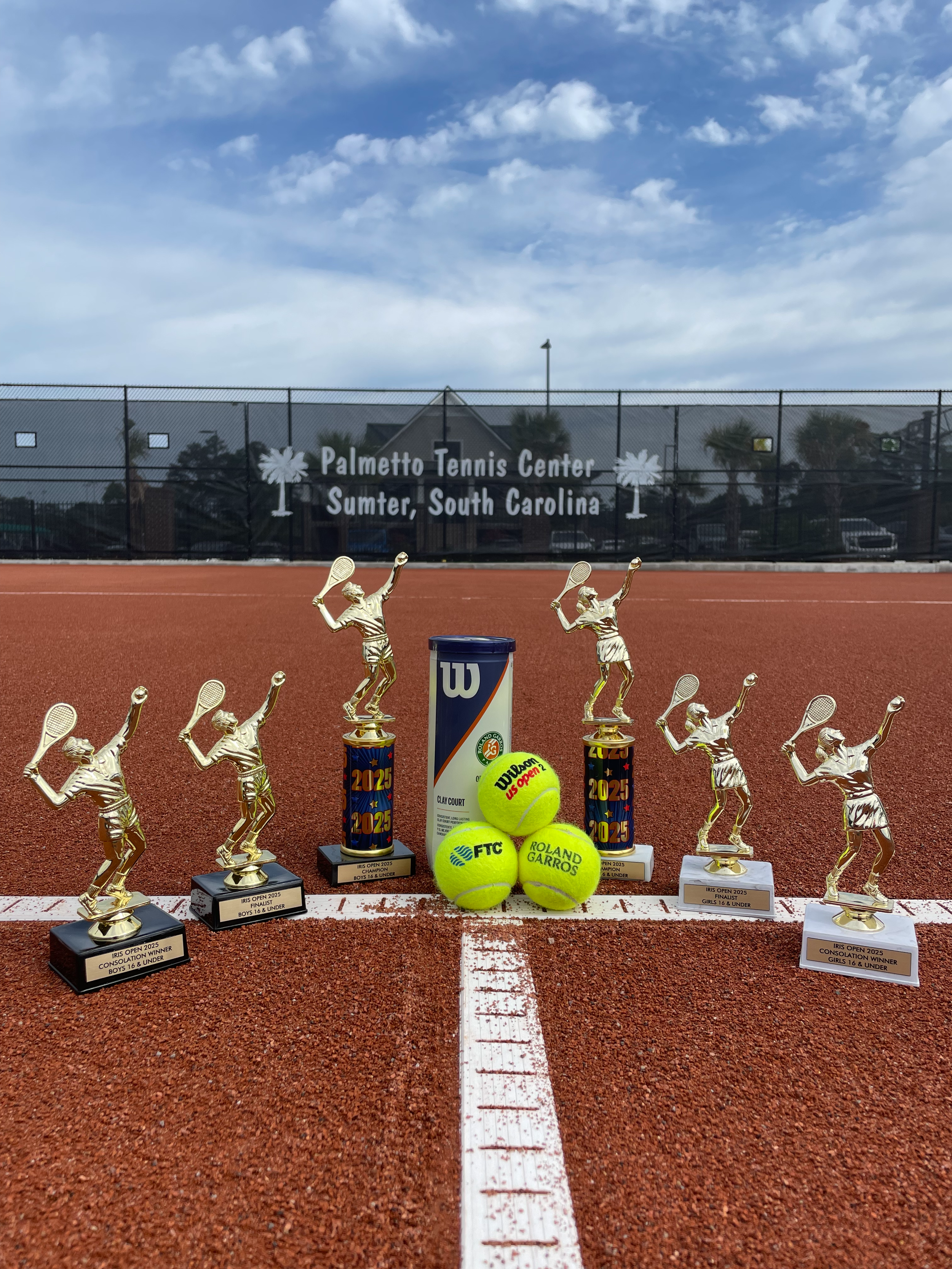 Tennis trophies, tennis balls, and tennis rackets displayed on a tennis court with a fence and a sign reading "Palmetto Tennis Center, Sumter, South Carolina" in the background.