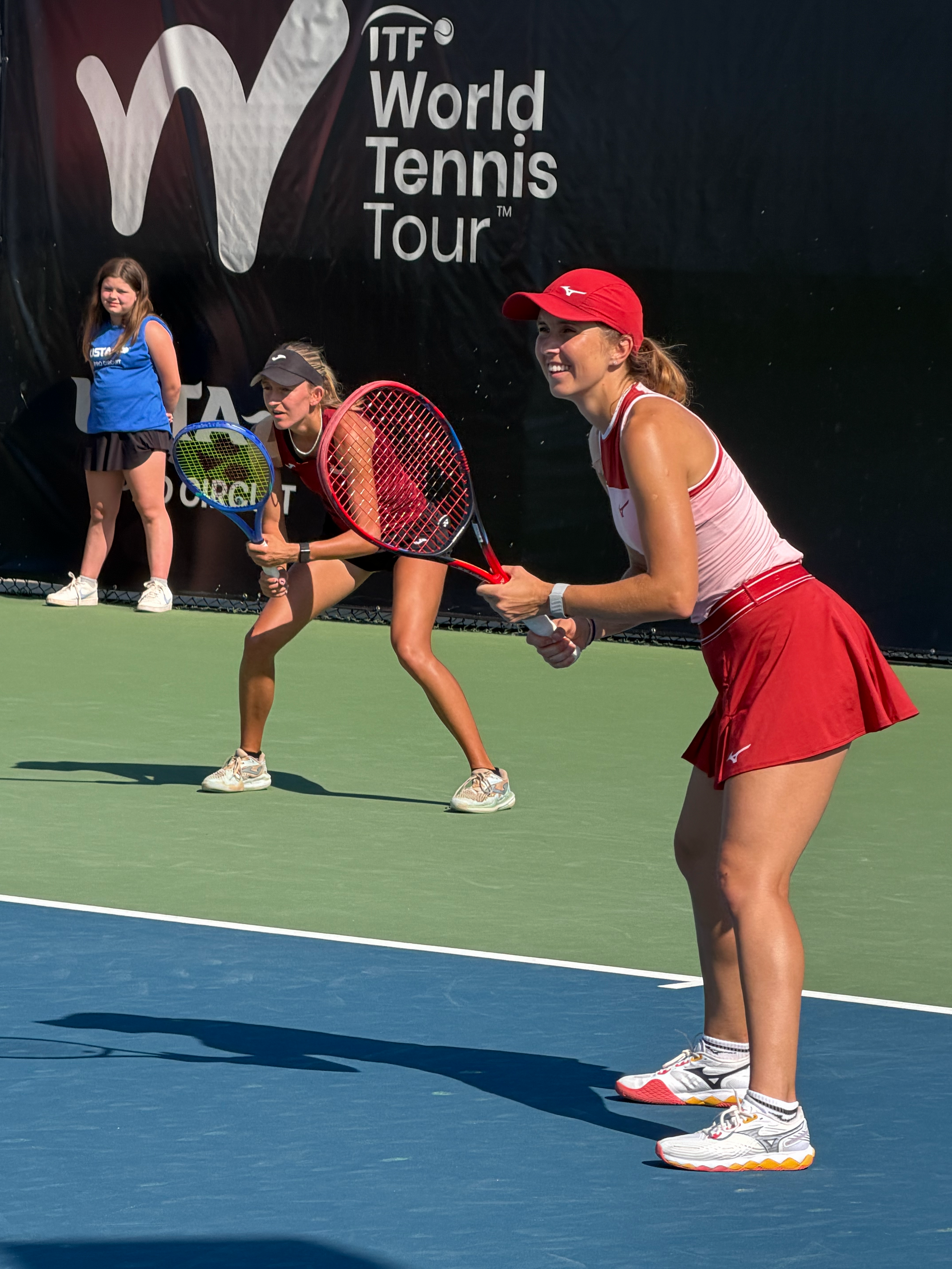 Three female tennis players in athletic clothing and sneakers on a tennis court preparing to hit a ball during a match at the ITF World Tennis Tour event.