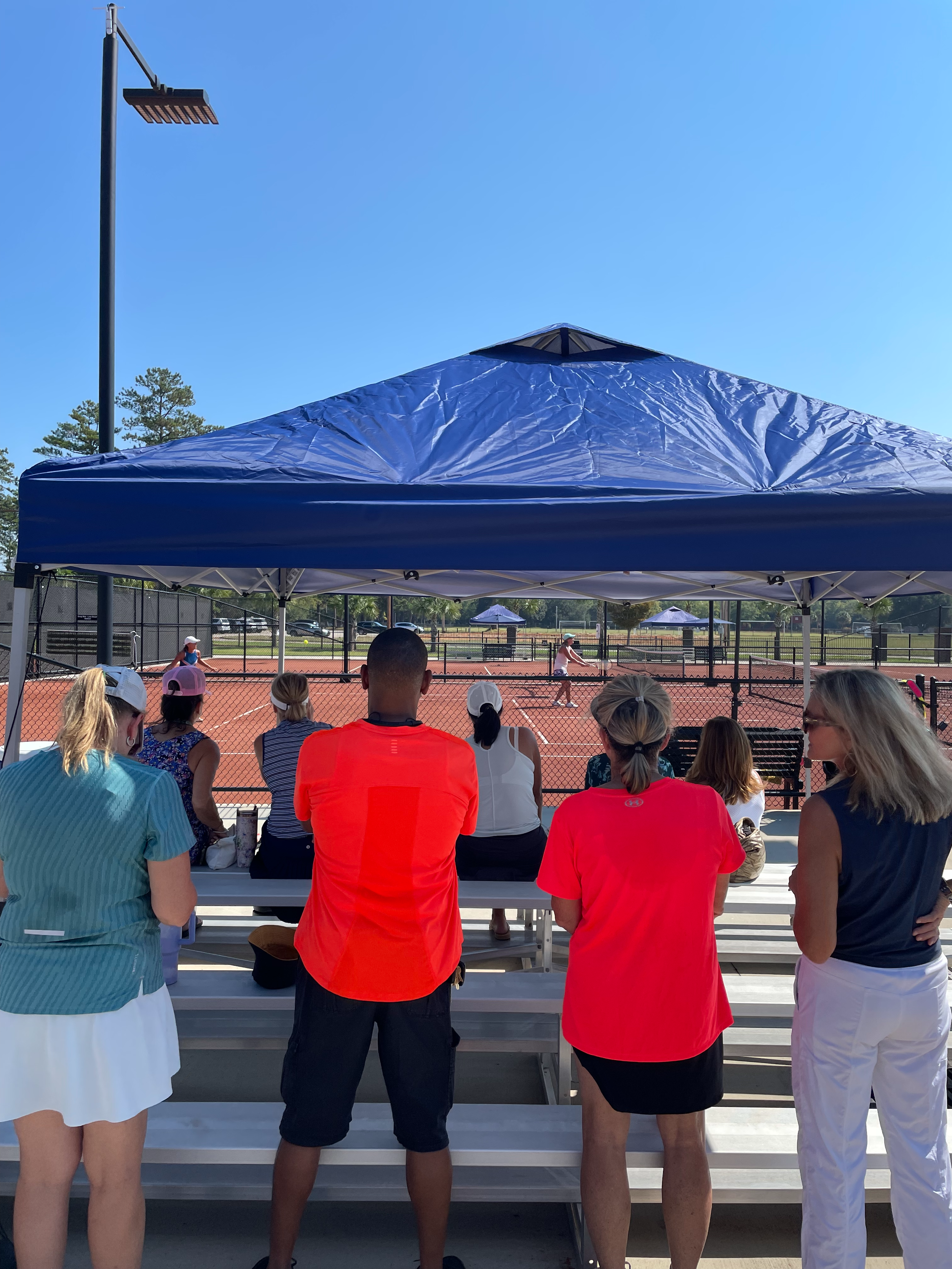 People sitting and standing outdoors, watching a tennis match under a blue canopy, with a tennis court in the background on a sunny day.