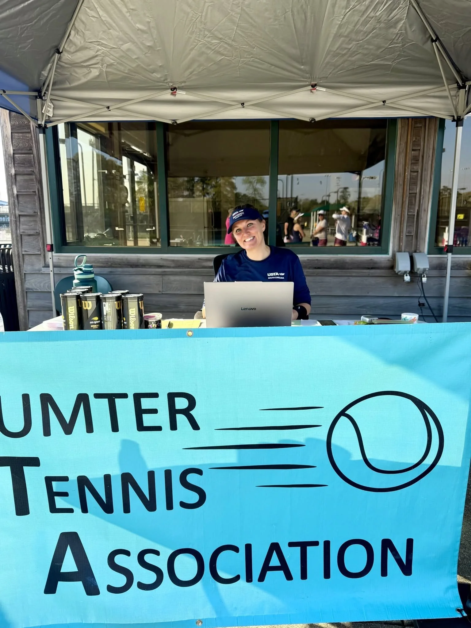 A woman wearing a navy blue athletic shirt and a cap sitting at a table with tennis balls and a laptop, with a large banner for Sumter tennis association in front.