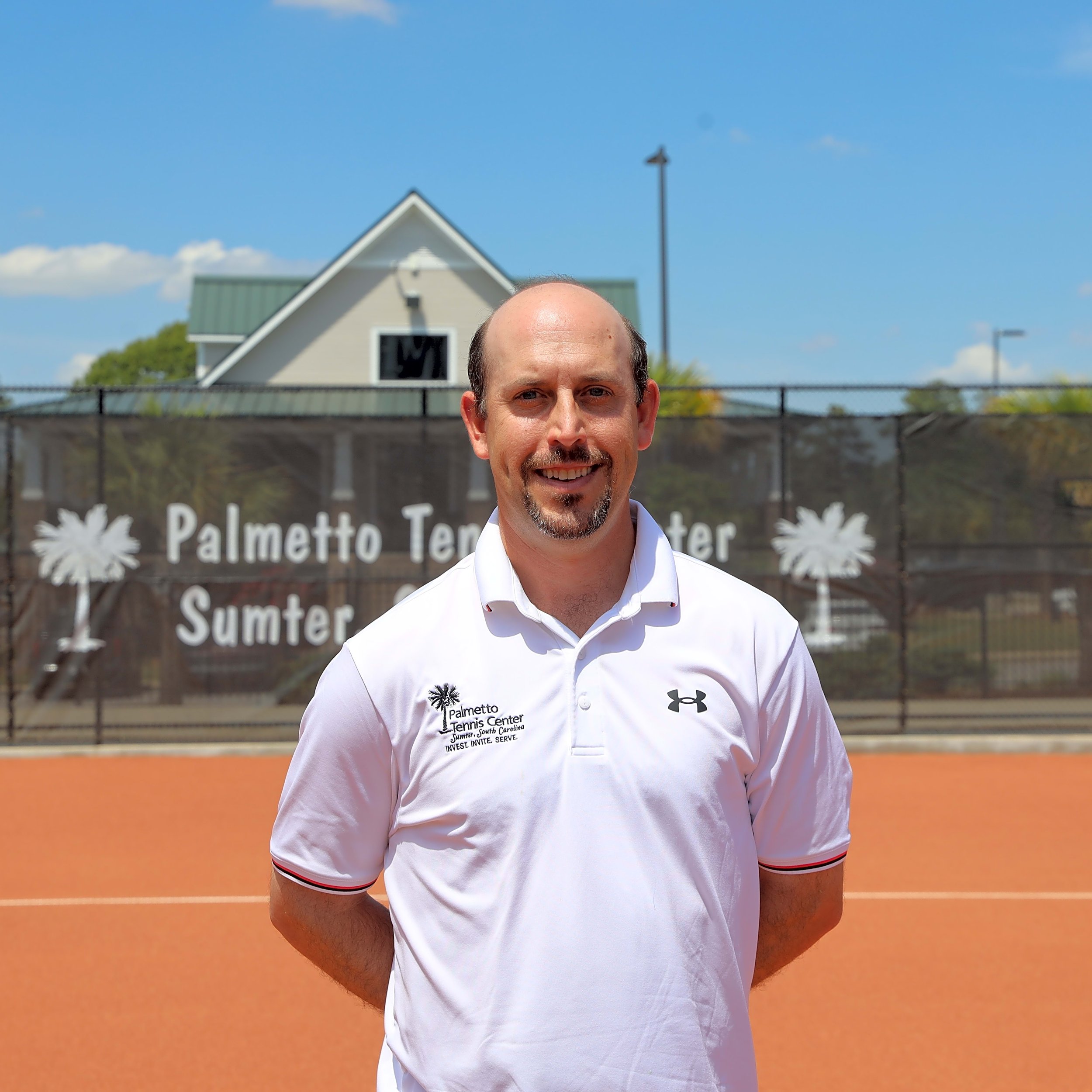 A man with a beard and mustache wearing a white collared shirt with Palmetto Tennis Center logo standing on a tennis court outdoors under blue sky.