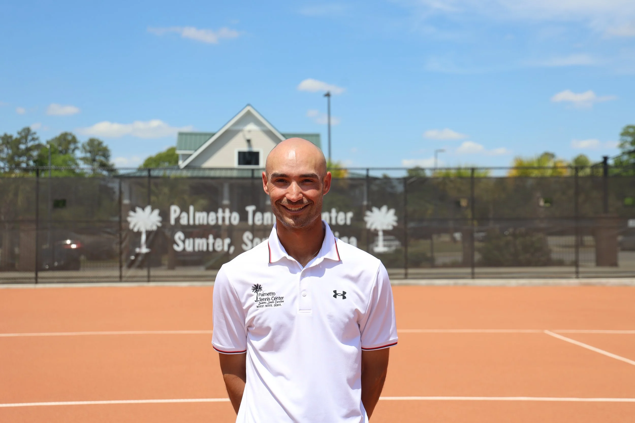 Smiling man in white shirt standing on a tennis court with a fence, trees, and a building in the background on a sunny day.