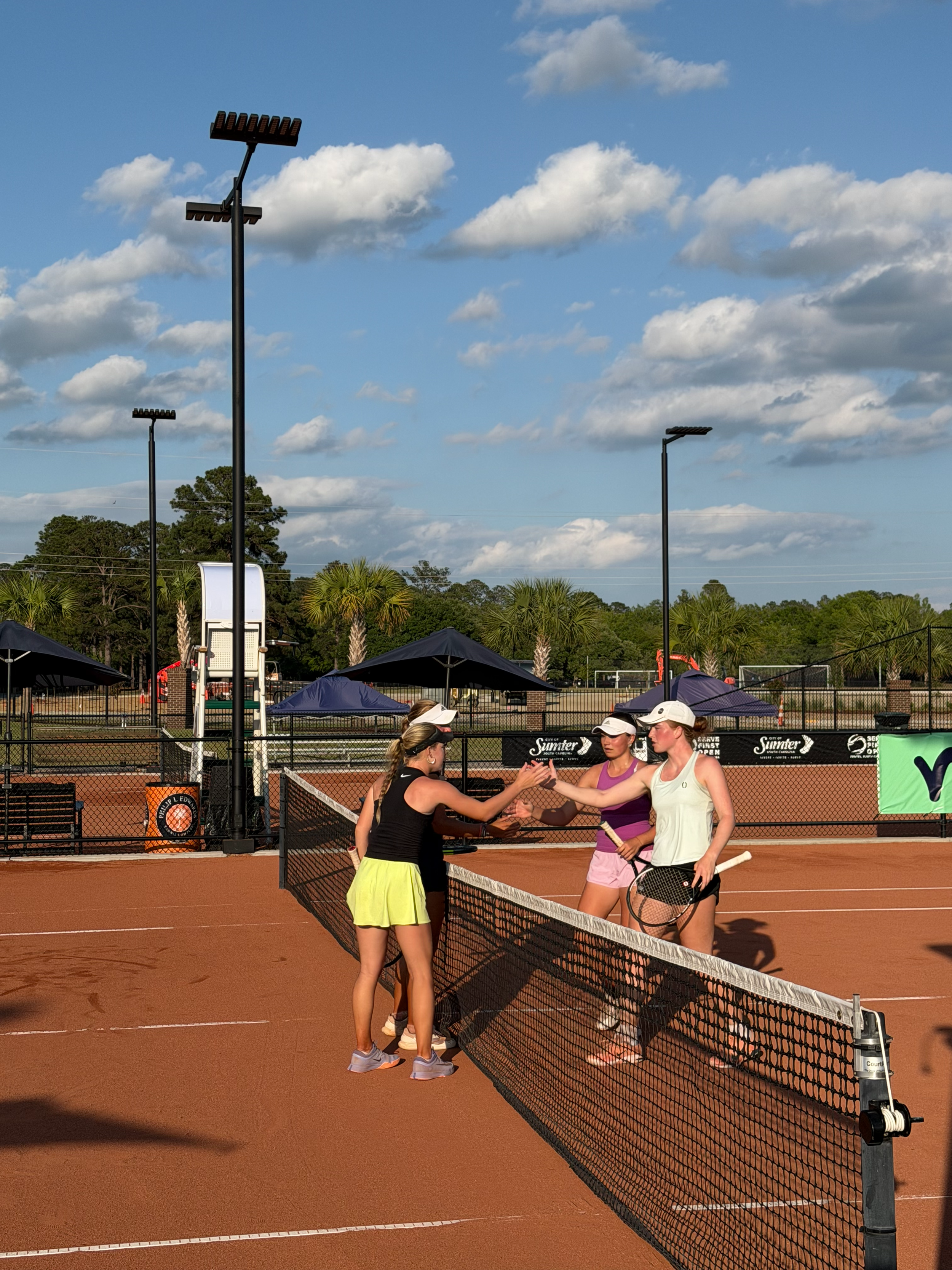 Four women are standing on a tennis court, shaking hands at the net after a match. They are dressed in athletic clothing with tennis rackets in hand. The tennis court is outdoors with a backdrop of trees and a blue sky with clouds.