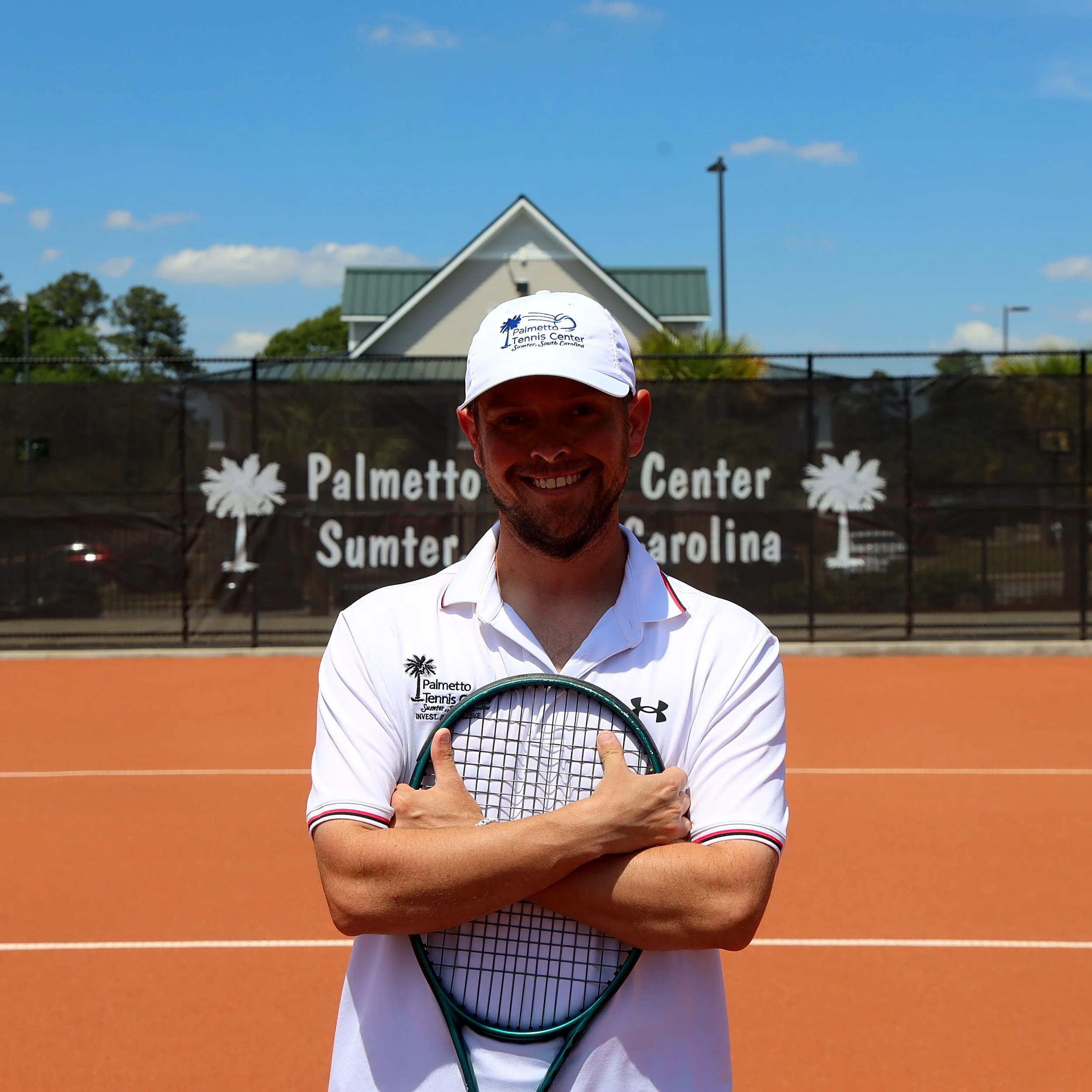 A man standing on a tennis court, smiling, wearing a white cap and a white shirt, holding a tennis racket in front of him. The background shows a sign for Palmetto Tennis Center in Summerville, South Carolina, with a building and a blue sky with some clouds.