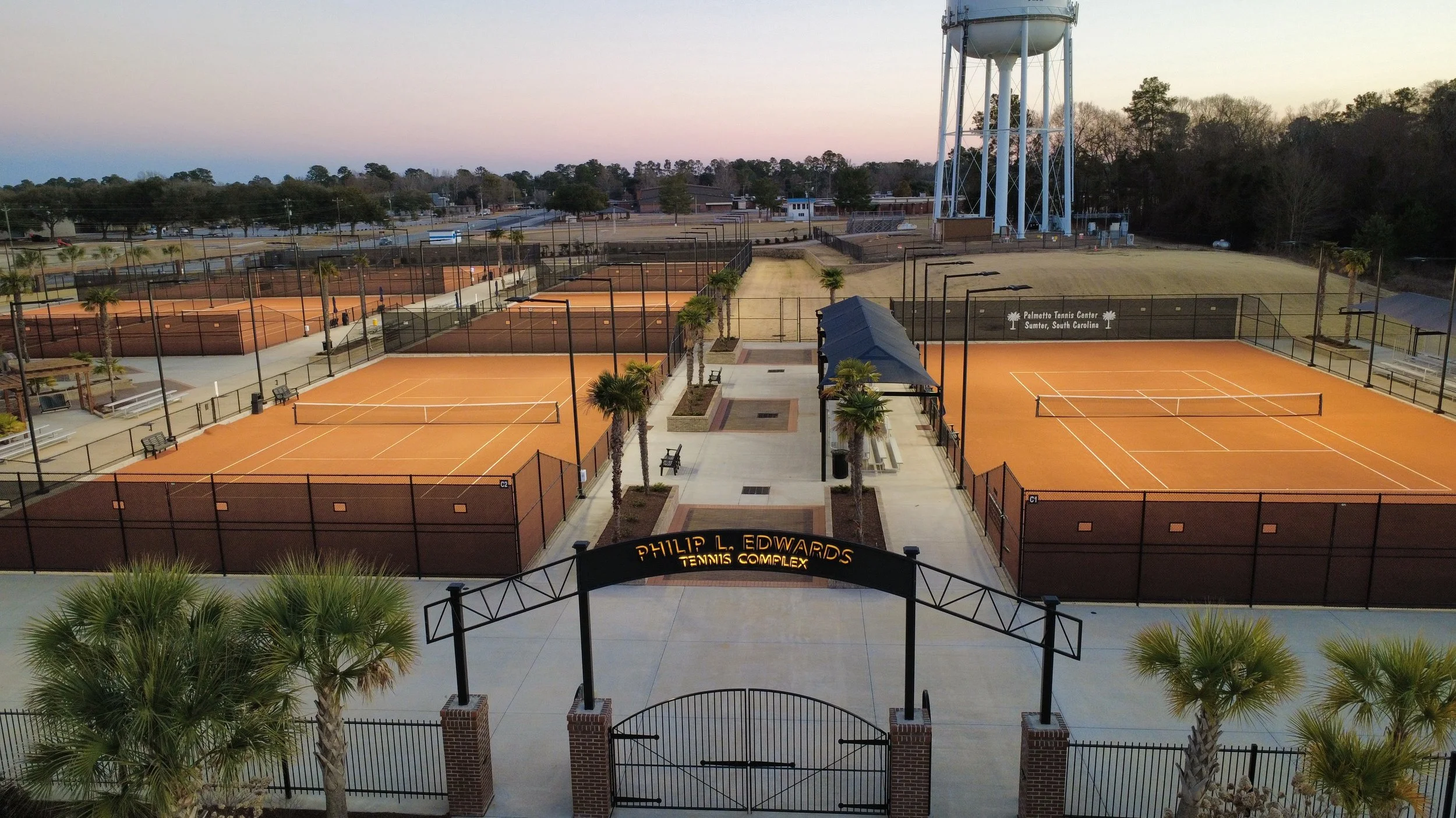 View of a tennis complex with multiple clay courts, surrounded by fencing and palm trees, at sunset.
