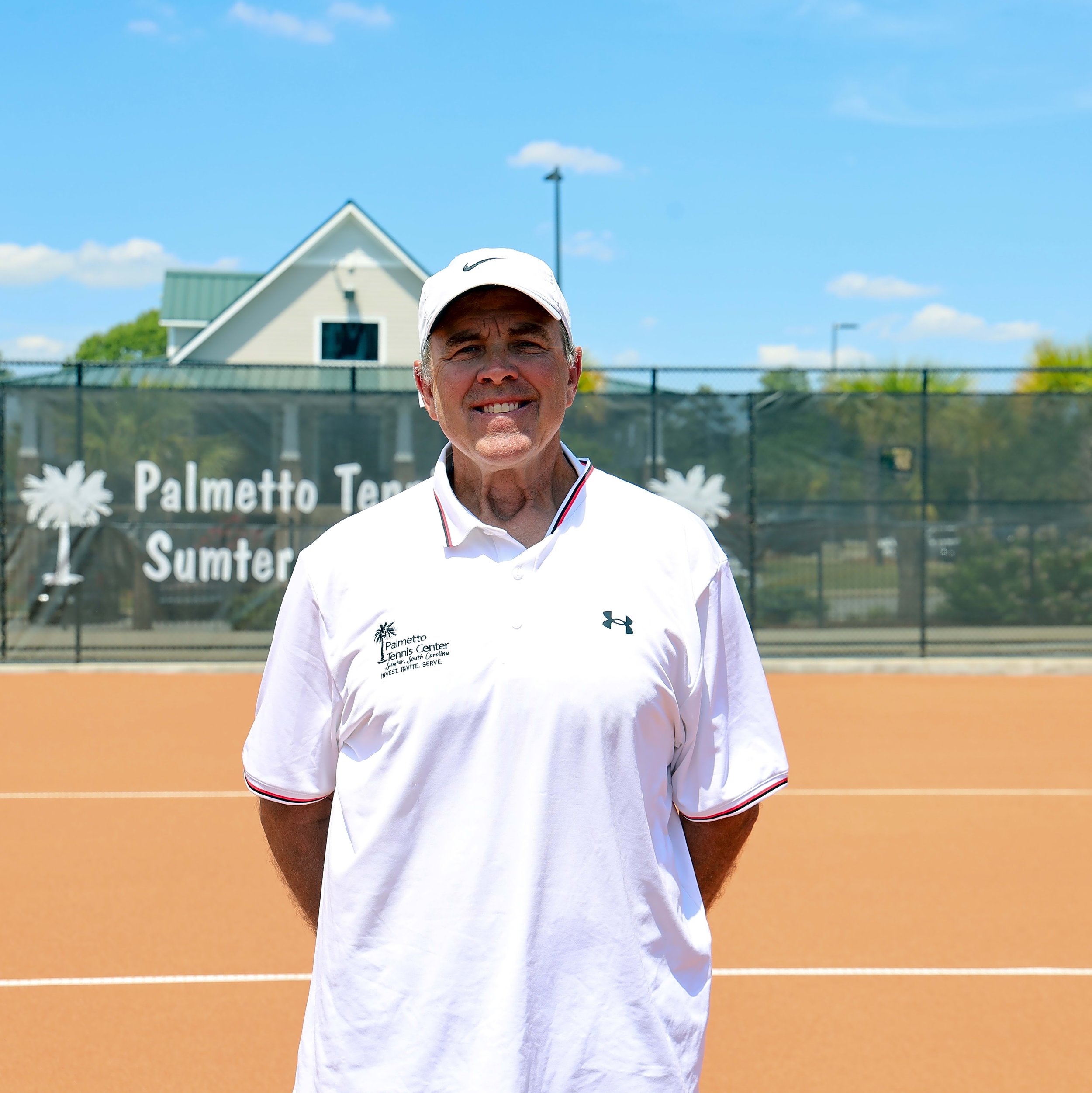 A man wearing a white Nike cap and white polo shirt at a tennis court with a sunny blue sky in the background.