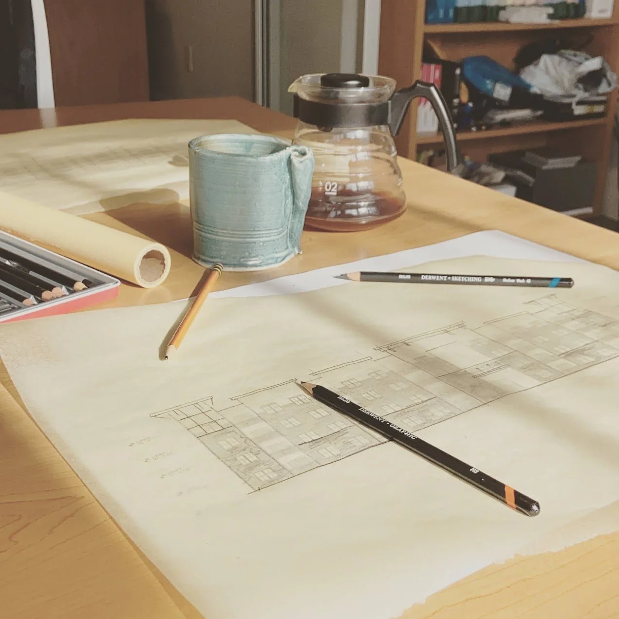 A workspace with architectural drawings, colored pencils, and sketching pens on a wooden table. In the background, there is a glass coffee pot, a blue ceramic mug, and a bookshelf.