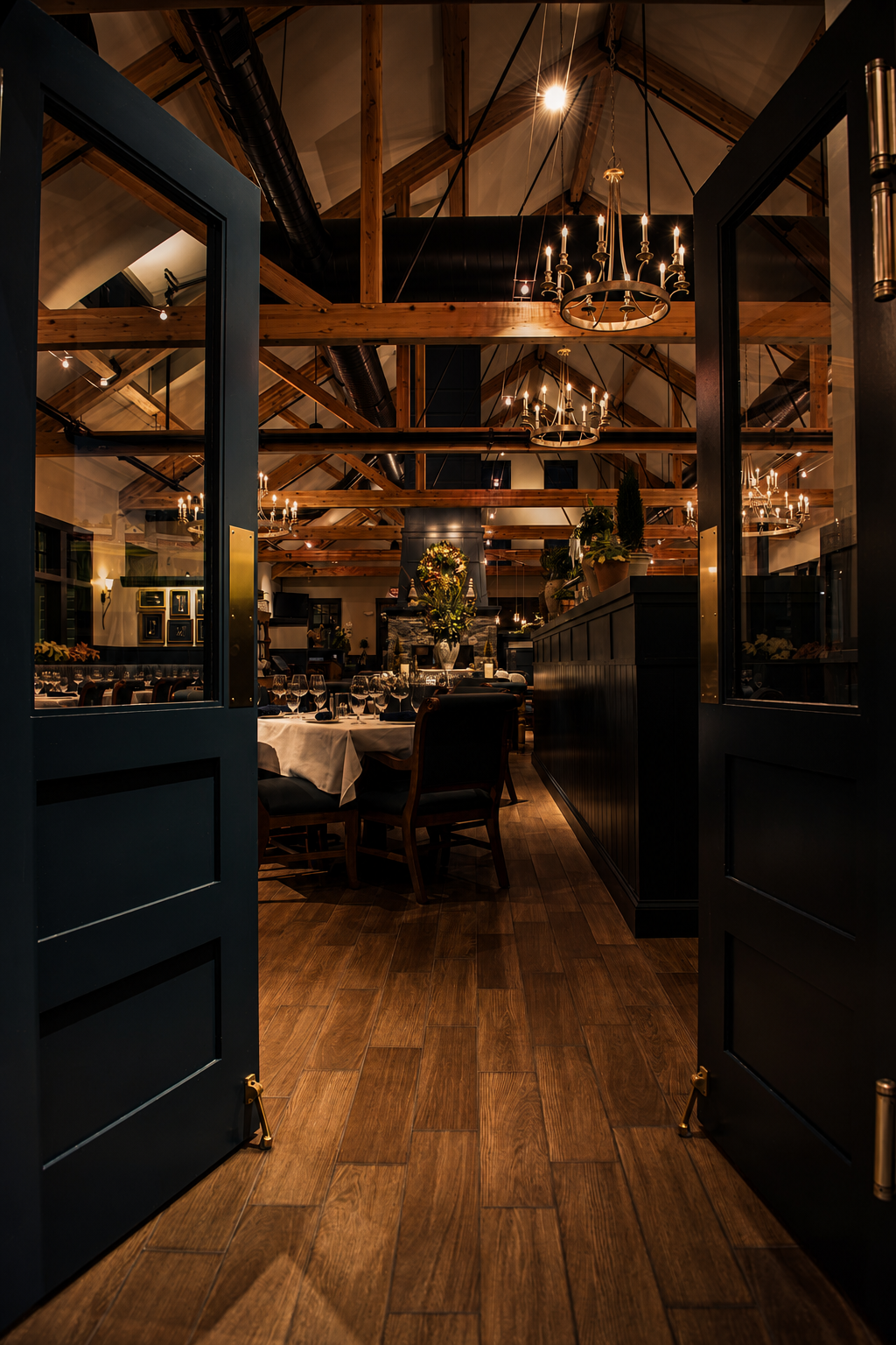 Elegant restaurant interior with wooden beams, chandeliers, and a dining table set with wine glasses and white tablecloths, viewed through open black doors.