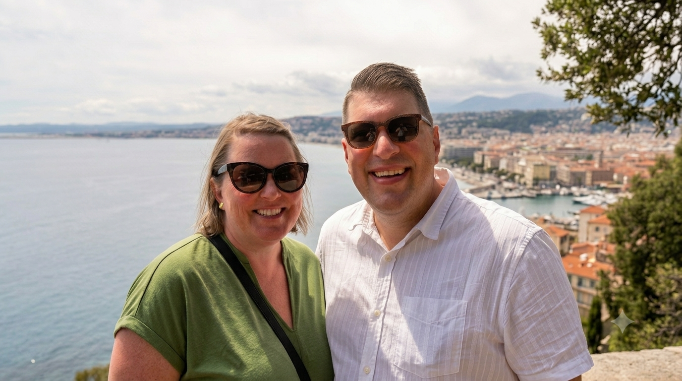 Two smiling people, a woman in a green shirt and a man in a white shirt, taking a selfie with a scenic coastal city and harbor in the background under cloudy skies.