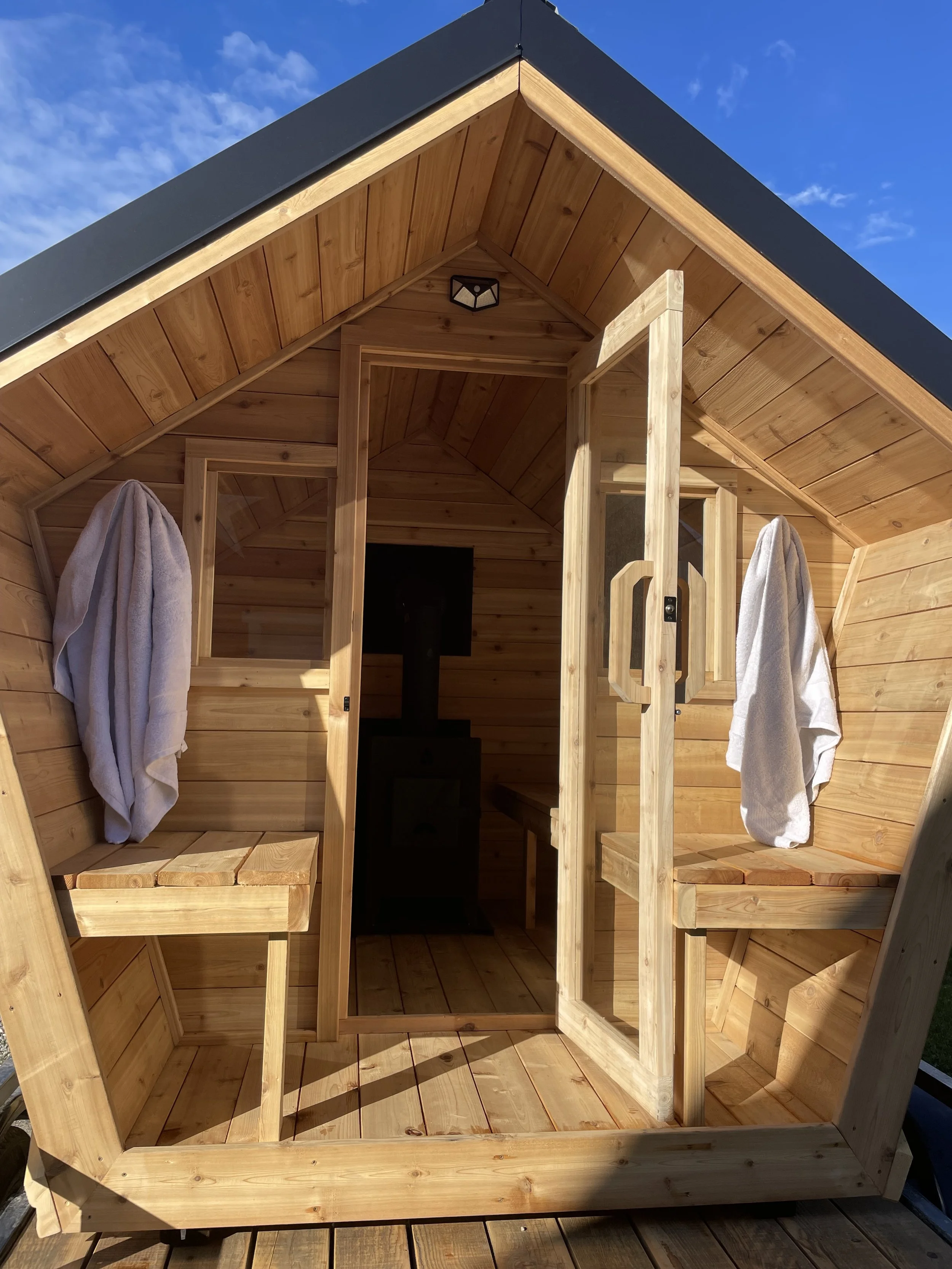 Inside a cedar sauna with benches on both sides, towels hanging on hooks, and a small window. The ceiling has wooden planks, and the outside sky is visible.
