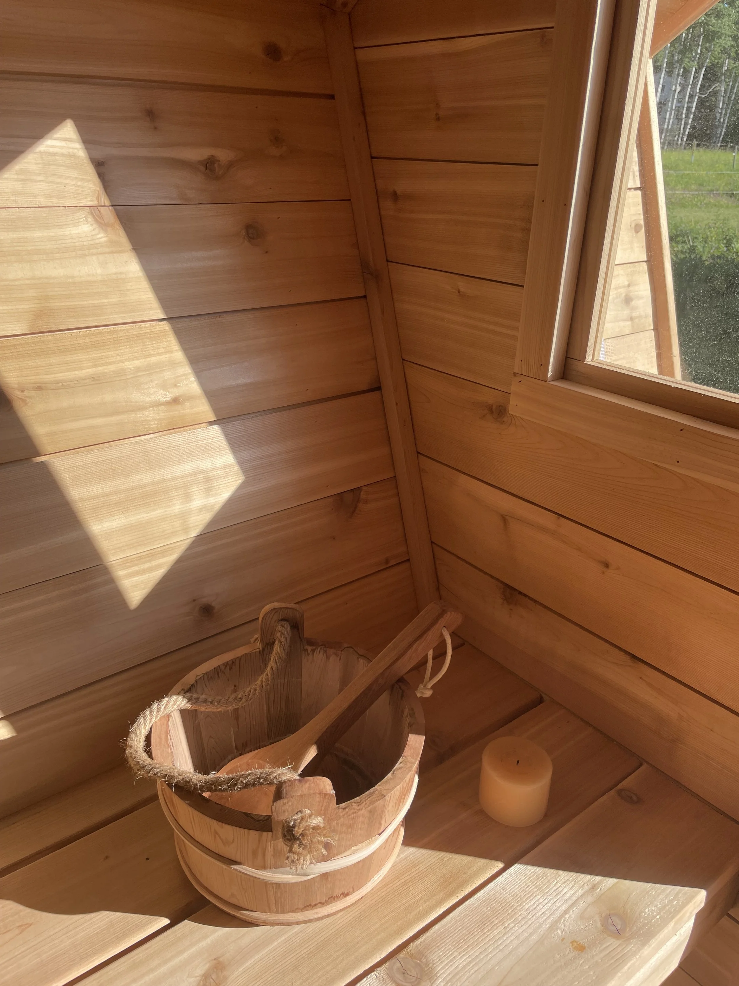 Cedar sauna interior with a bucket, a candle, and a window showing greenery outside.