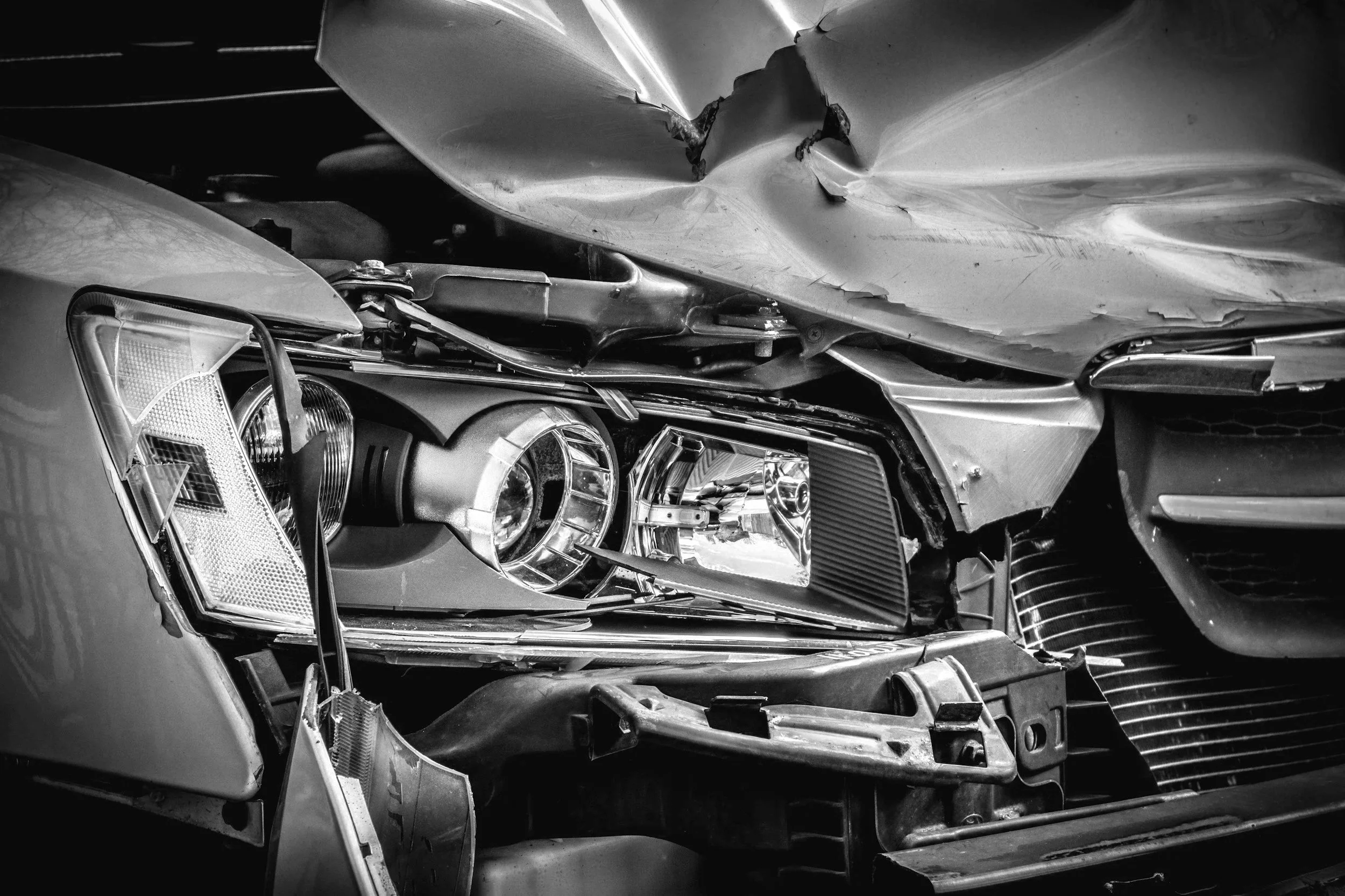 Black and white photo of a damaged car front end, showing a crumpled hood, broken headlight, and bent grille