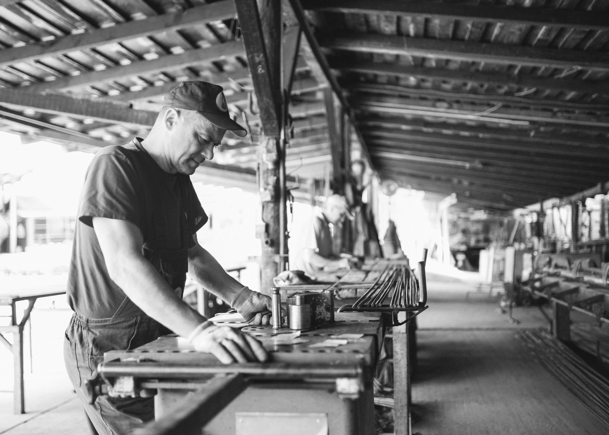 A man working with woodworking tools in a workshop with a long table and a roof structure. Black and white photo.