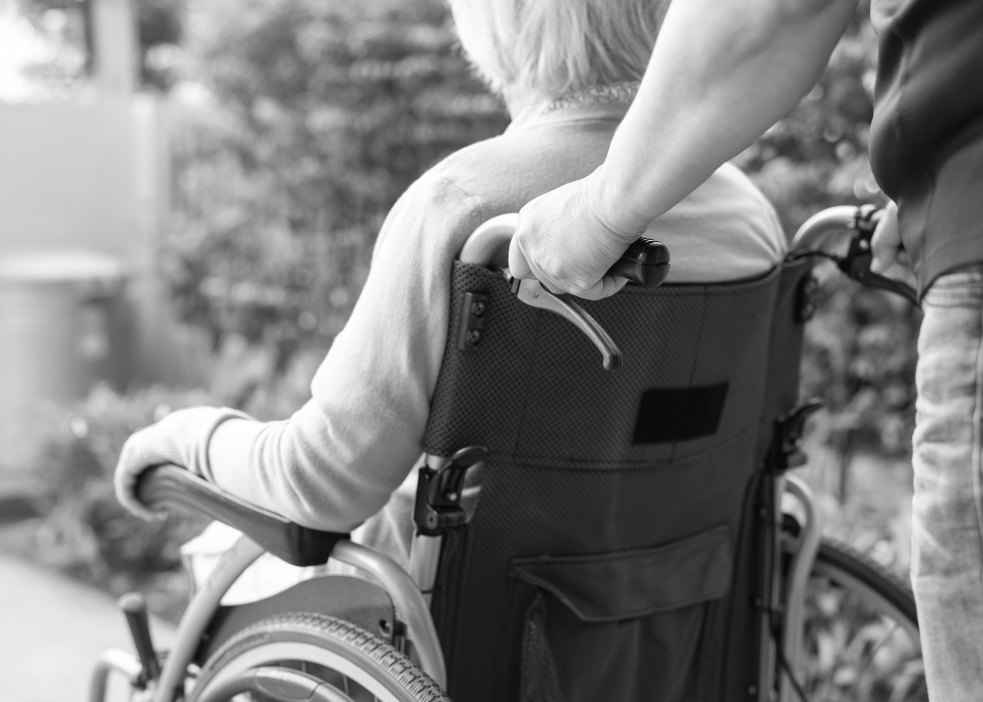 Close-up black and white photo of a person in a wheelchair holding the wheel's handle in an outdoor setting with blurred trees in the background.